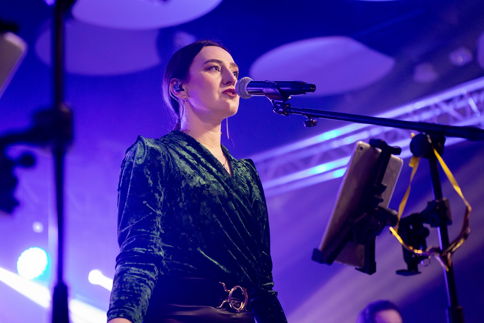 Wokalistka zespołu muzycznego z Poznania, w aksamitnej, zielonej bluzce, śpiewa do mikrofonu podczas występu na scenie oświetlonej fioletowym światłem, z tabletem umieszczonym na statywie obok