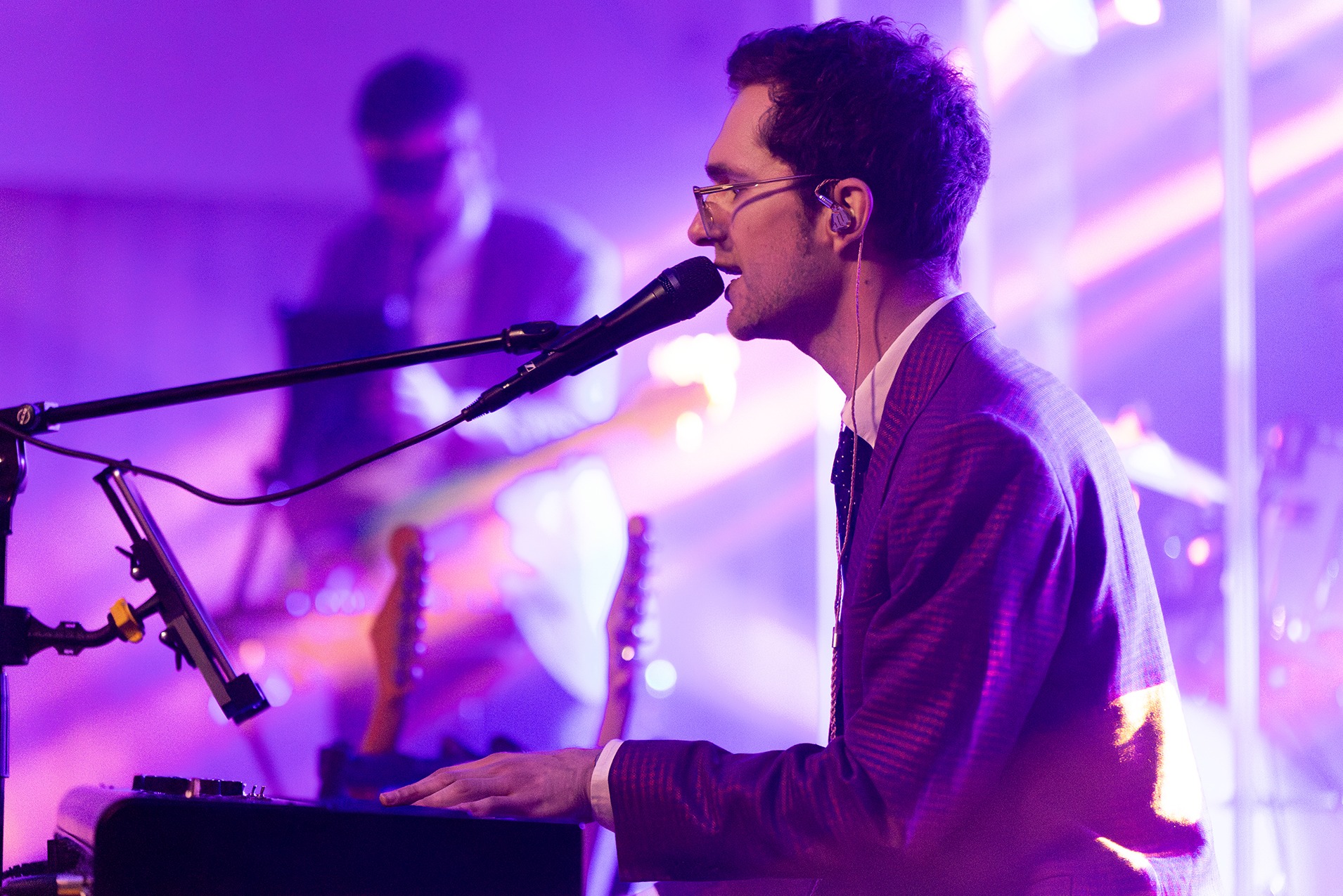 Mężczyzna w okularach i garniturze gra na keyboardzie, śpiewając do mikrofonu podczas występu na scenie z gitarzystą w tle, oświetlonej fioletowymi światłami.