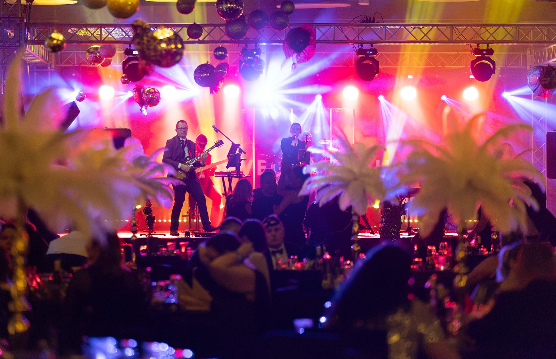 Scena z muzykami podczas występu na tle dekoracji z kulami dyskotekowymi, widziana zza stolików gości.