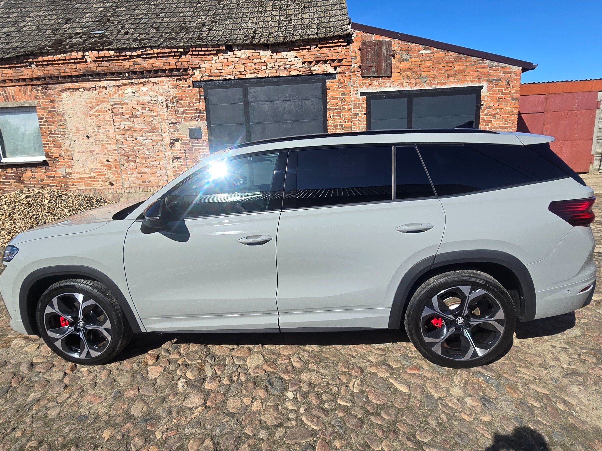 Szary SUV z przyciemnionymi szybami, zaparkowany na brukowanej nawierzchni przed ceglanym budynkiem w słoneczny dzień. Widoczne czerwone zaciski hamulcowe.