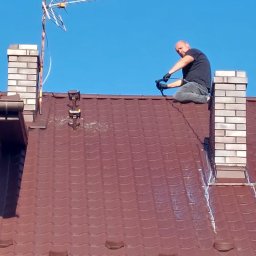 INSECT s. c. - Pracownik w czarnych rękawicach na kolanach na brązowym dachu z kominem, instalujący element przy antenie telewizyjnej na tle błękitnego nieba.