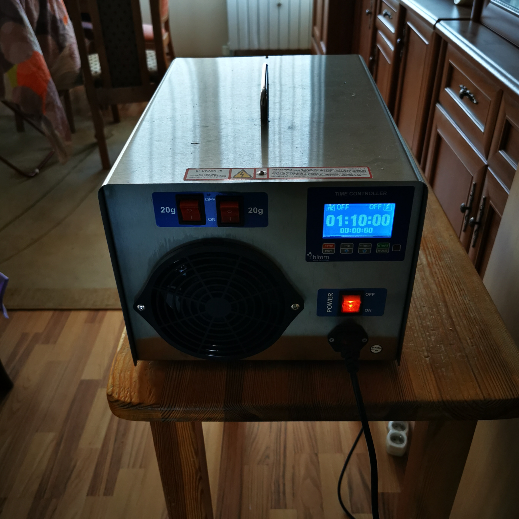 Generator ozonu Bitom z wyświetlaczem czasu pracy i przełącznikami, ustawiony na drewnianym stoliku, z widocznym kablem zasilającym i gniazdkiem.