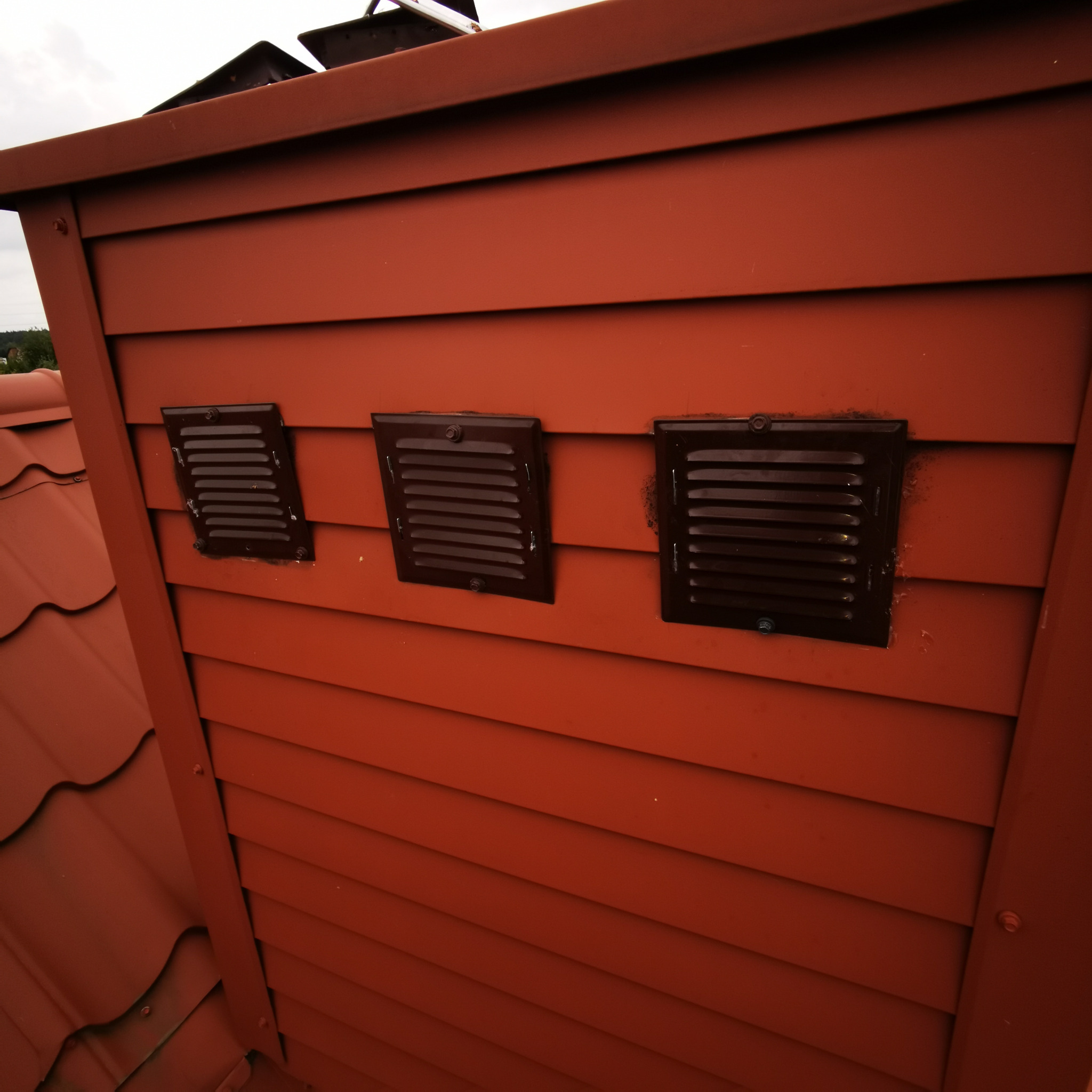Czerwona drewniana obudowa komina z trzema brązowymi kratkami wentylacyjnymi, widoczne elementy dachu z dachówki ceramicznej.
