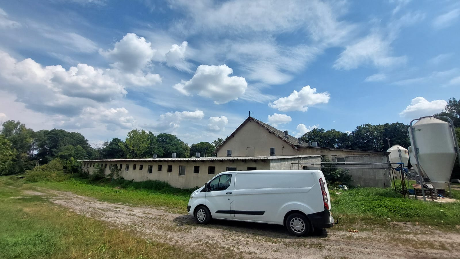 Biały van zaparkowany na polnej drodze przed budynkiem gospodarczym z licznymi oknami i silosami w tle, pod błękitnym niebem z obłokami.