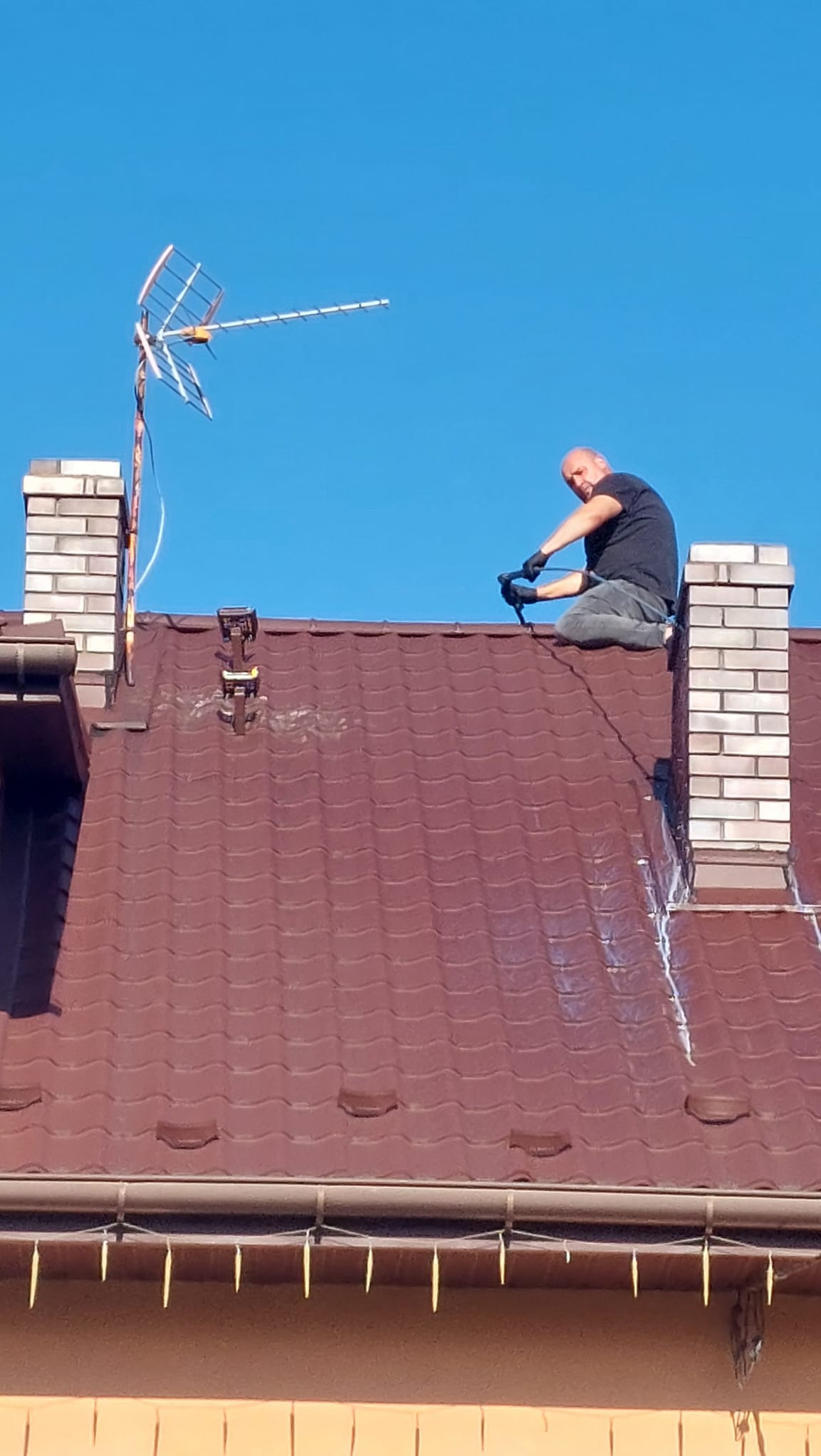 Pracownik w czarnych rękawicach na kolanach na brązowym dachu z kominem, instalujący element przy antenie telewizyjnej na tle błękitnego nieba.