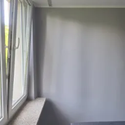 Jasnoszara ściana po malowaniu, widoczne okno z parapetem w stylu lastryko i fragment sofy. Minimalistyczne wnętrze w chłodnej tonacji.