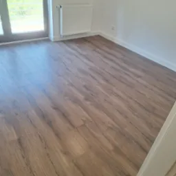 Jasne panele podłogowe z widoczną strukturą drewna w pustym pokoju z białymi ścianami i grzejnikiem. Okno balkonowe z widokiem na zieleń.