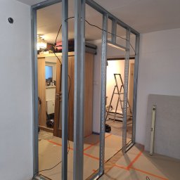 Ivan Katrych - Metalowa konstrukcja ścianki działowej w trakcie montażu, widoczne profile i kable. Na podłodze taśma wyznaczająca obszar robót. W tle drabina i płyta gipsowa.