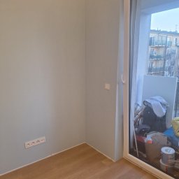 Ivan Katrych - Jasne, świeżo pomalowane wnętrze z drewnianą podłogą i widokiem na balkon przez duże okno. Widoczne gniazdka elektryczne i puszka farby.