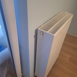 Ivan Katrych - Biały grzejnik panelowy zamontowany przy pomalowanej na jasny błękit ścianie, obok białej ramy okiennej i drewnianej podłogi. Ujęcie z dołu.