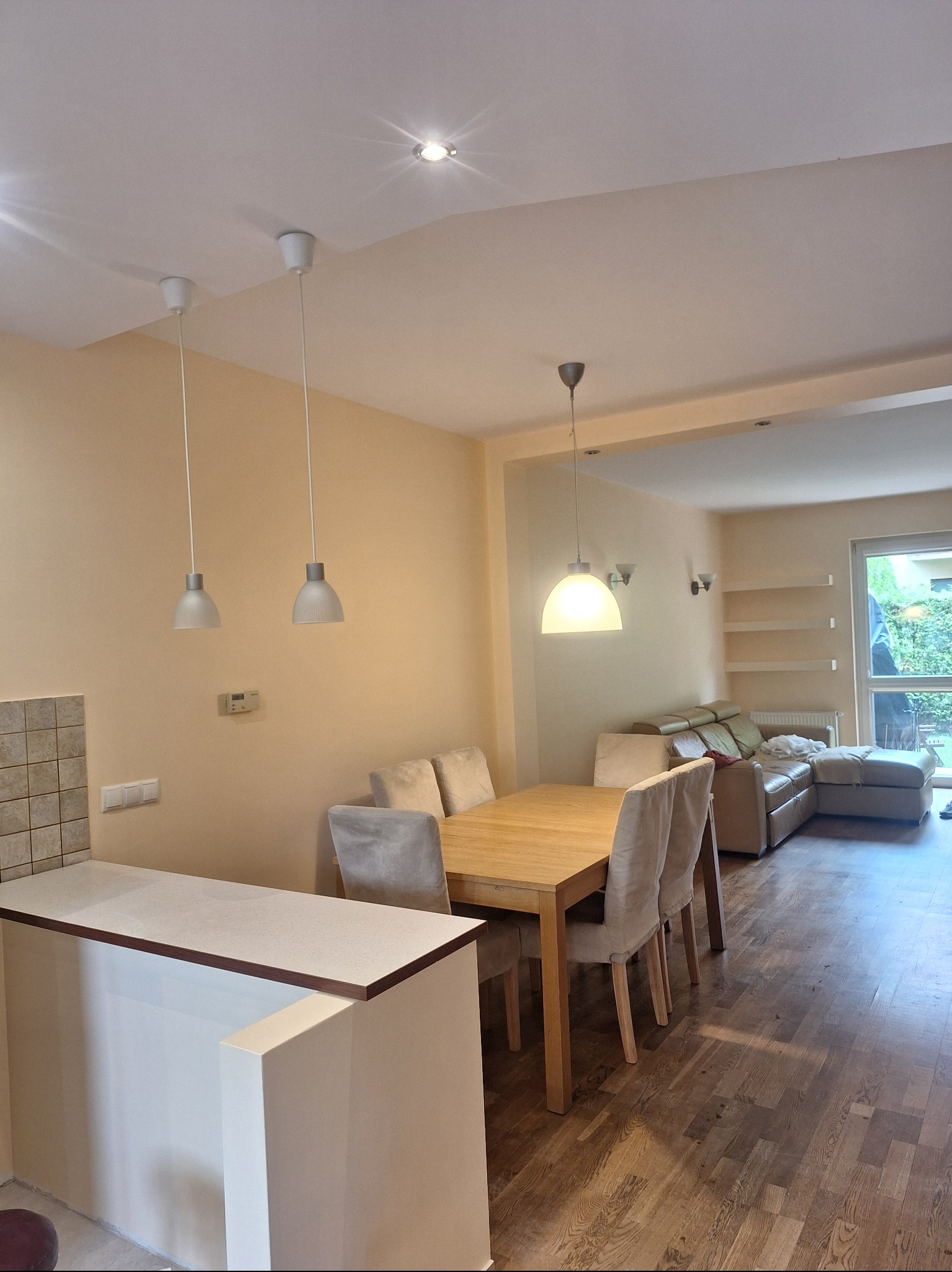 Jasne wnętrze po odświeżeniu: drewniana podłoga, stół z krzesłami, beżowa sofa i ściany w ciepłym odcieniu. Nowoczesne lampy wiszące dodają subtelnego charakteru.