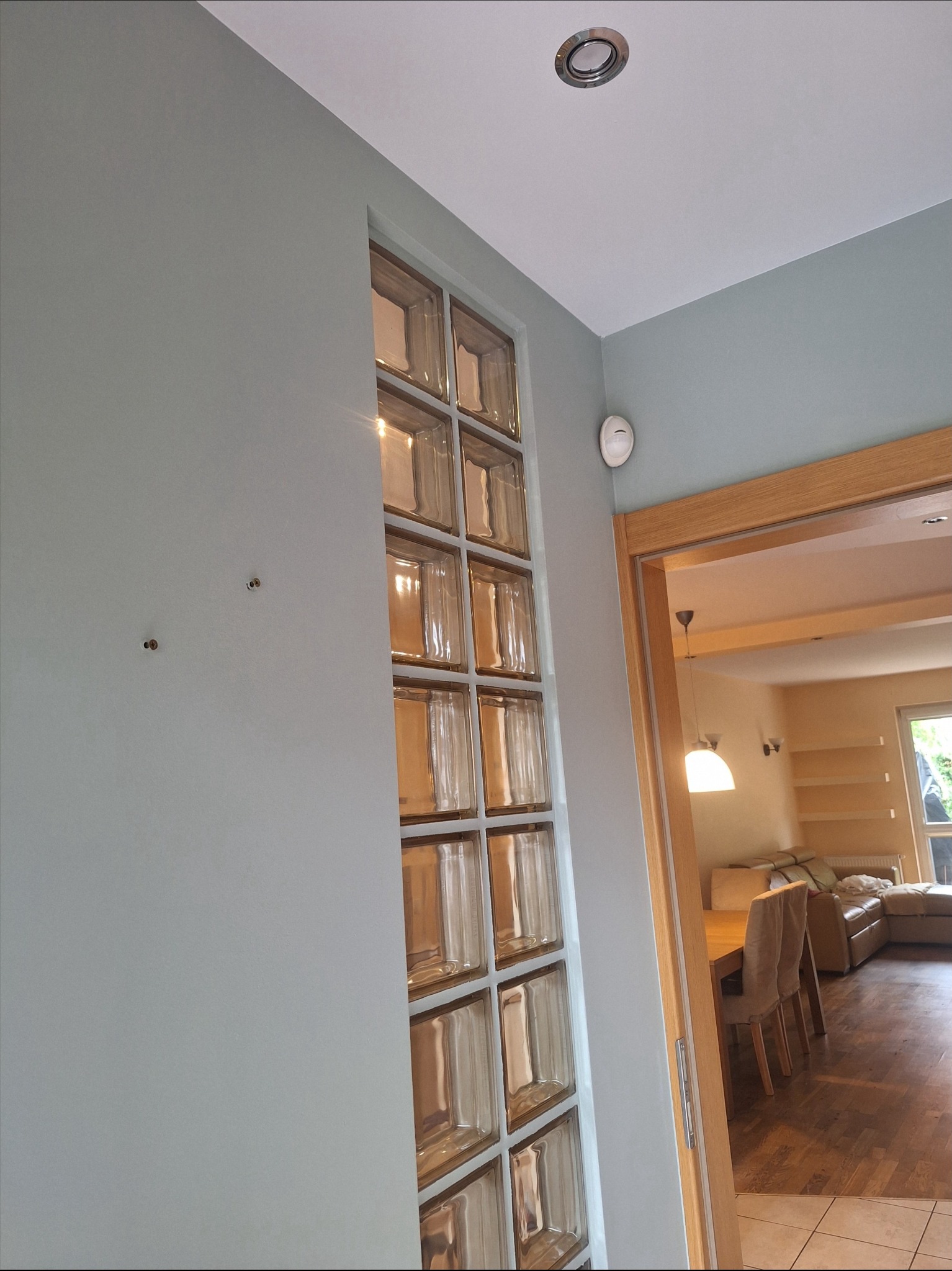 Ściana w odcieniach szarości z wbudowaną ścianką z luksferów, widoczny fragment pokoju z drewnianą podłogą i meblami. Oświetlenie sufitowe.