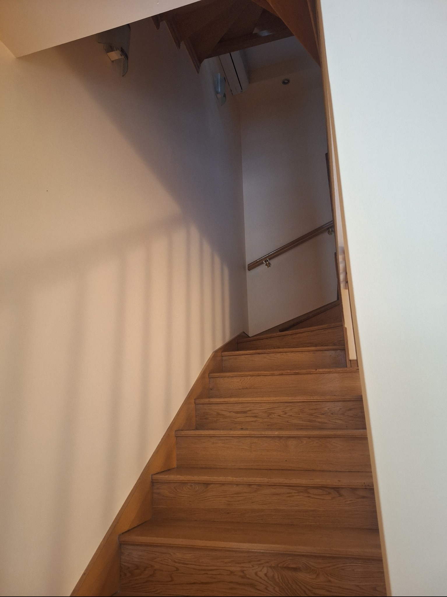 Drewniane schody wewnętrzne z poręczą, prowadzące na górę w jasnym wnętrzu. Widoczne detale wykończenia i oświetlenie.