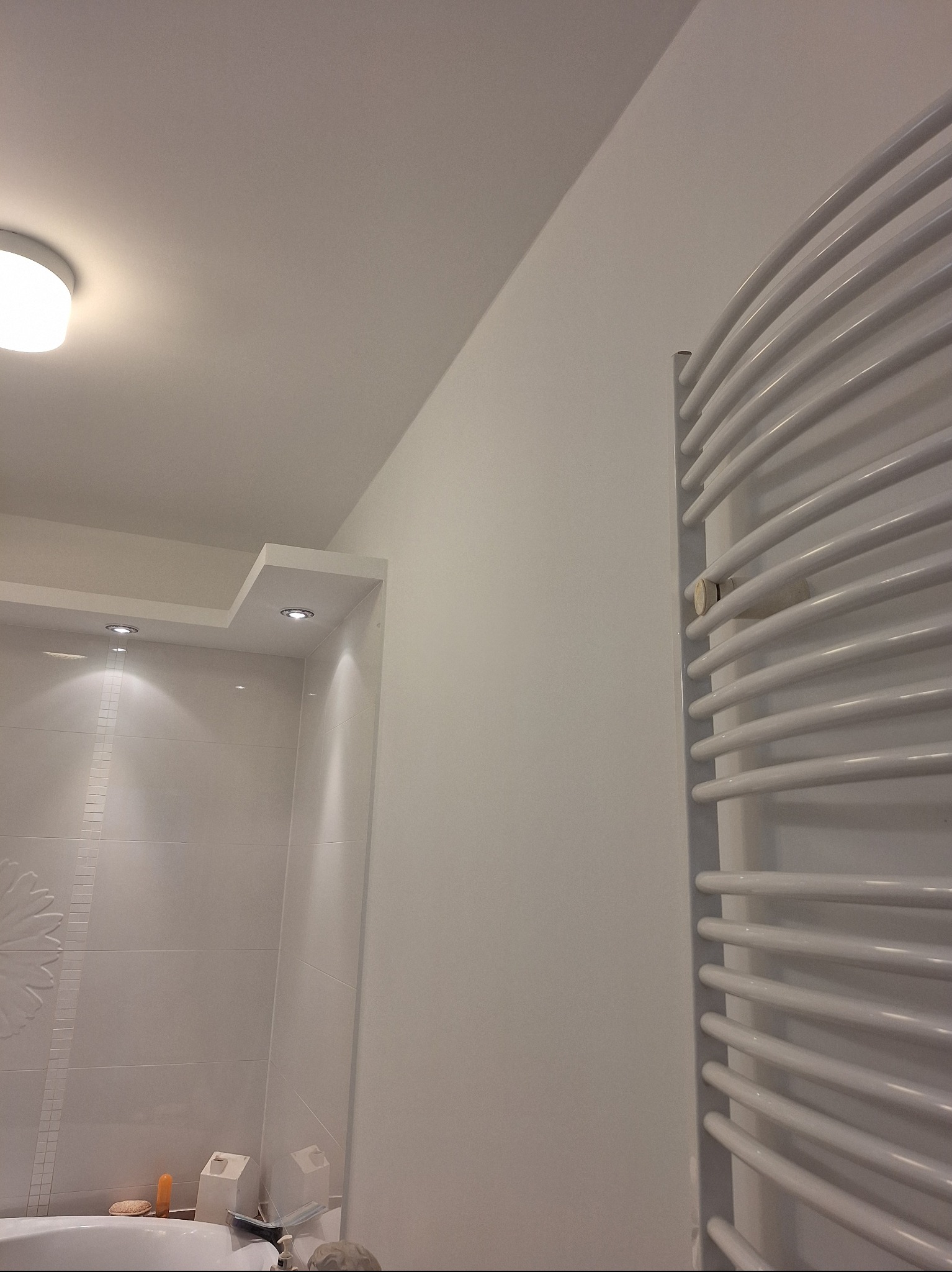 Minimalistyczna łazienka w bieli: grzejnik drabinkowy, oświetlenie punktowe nad wanną z dekoracyjną mozaiką. Nowoczesne wykończenie wnętrz.