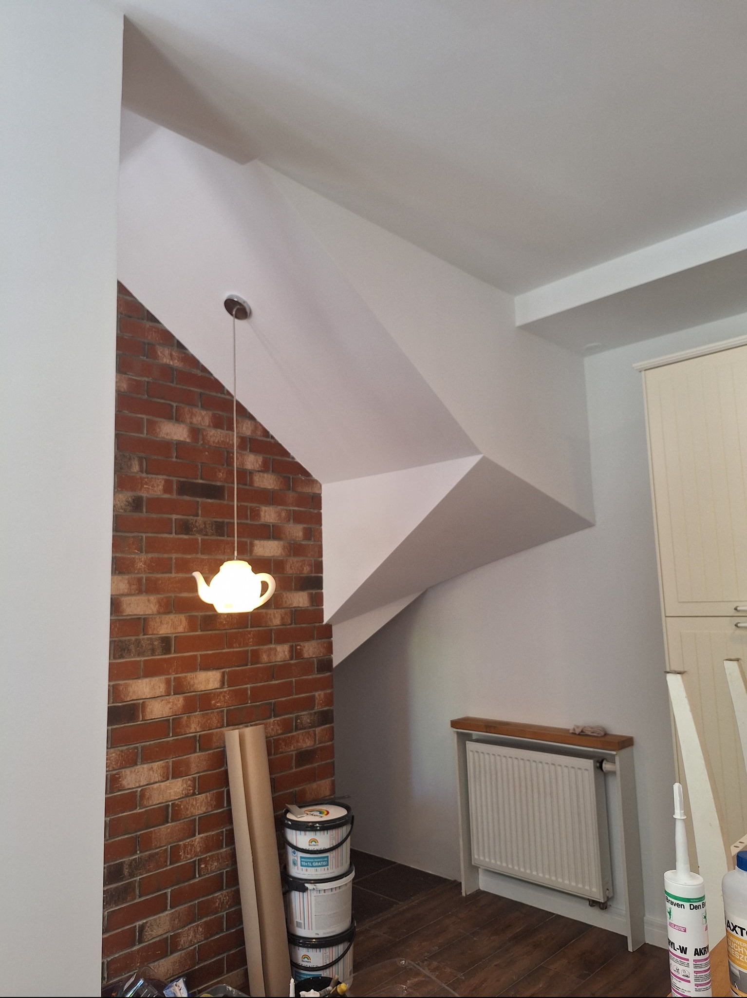 Wnętrze po odświeżeniu: białe ściany, ceglana ściana akcentująca z lampą w kształcie czajnika. Widoczne farby i akcesoria malarskie, grzejnik. Ukończony etap prac.