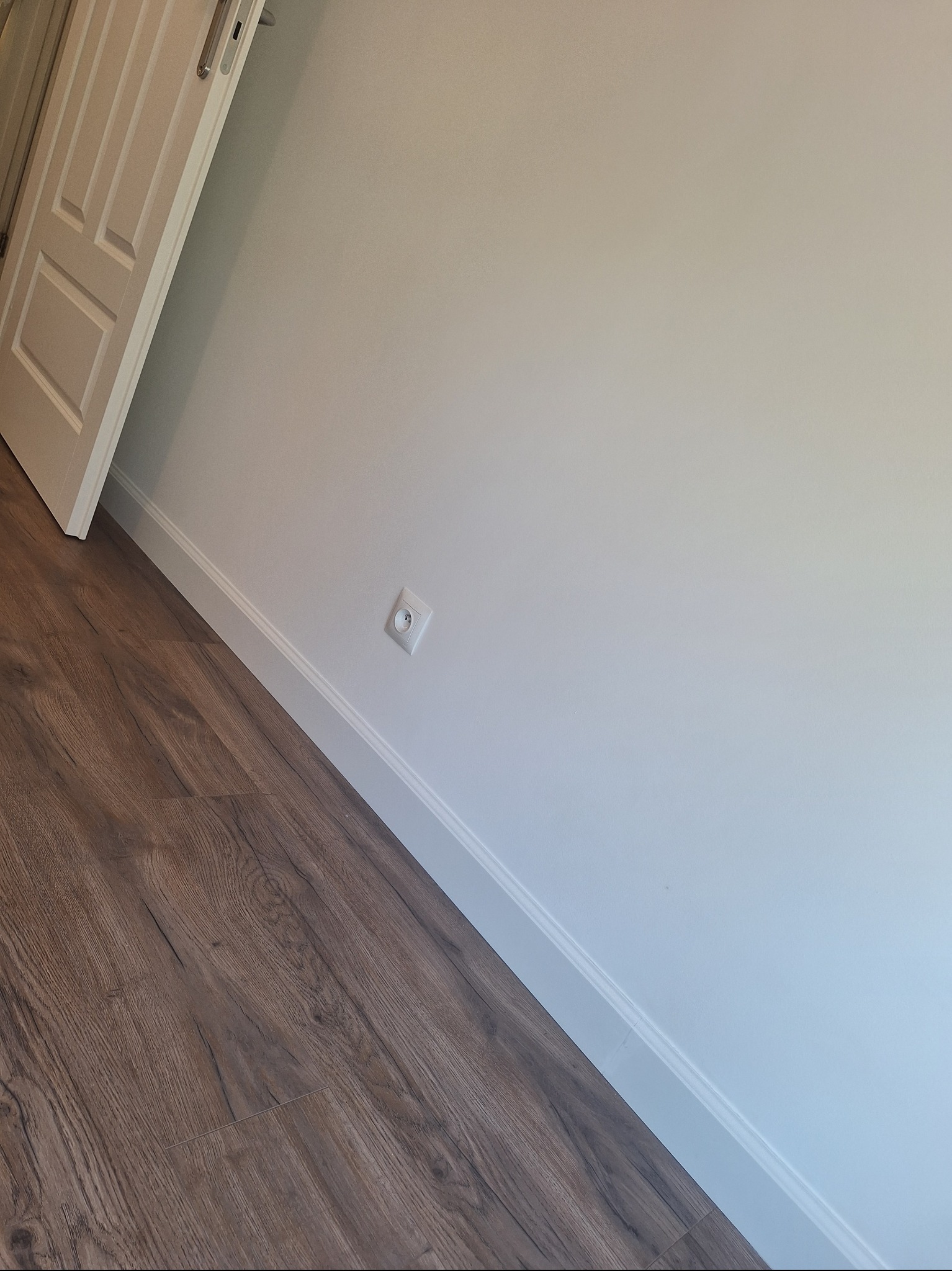 Ukończone wnętrze: brązowe panele podłogowe, biała ściana z listwą przypodłogową i gniazdkiem elektrycznym, białe drzwi z srebrną klamką.