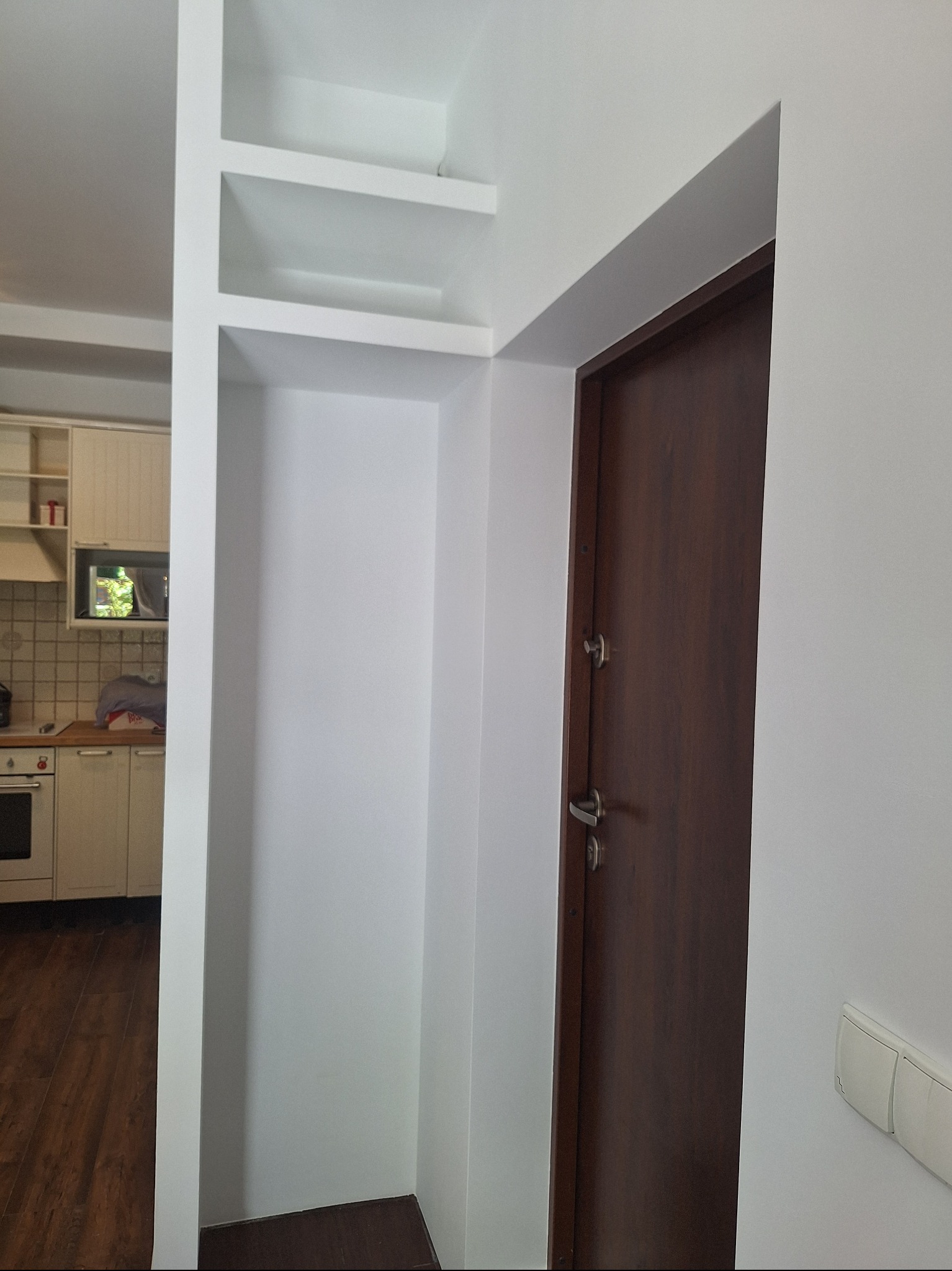 Wnęka z półkami pomalowana na biało, widoczny fragment kuchni i brązowe drzwi wejściowe. Minimalistyczne wykończenie wnętrza.