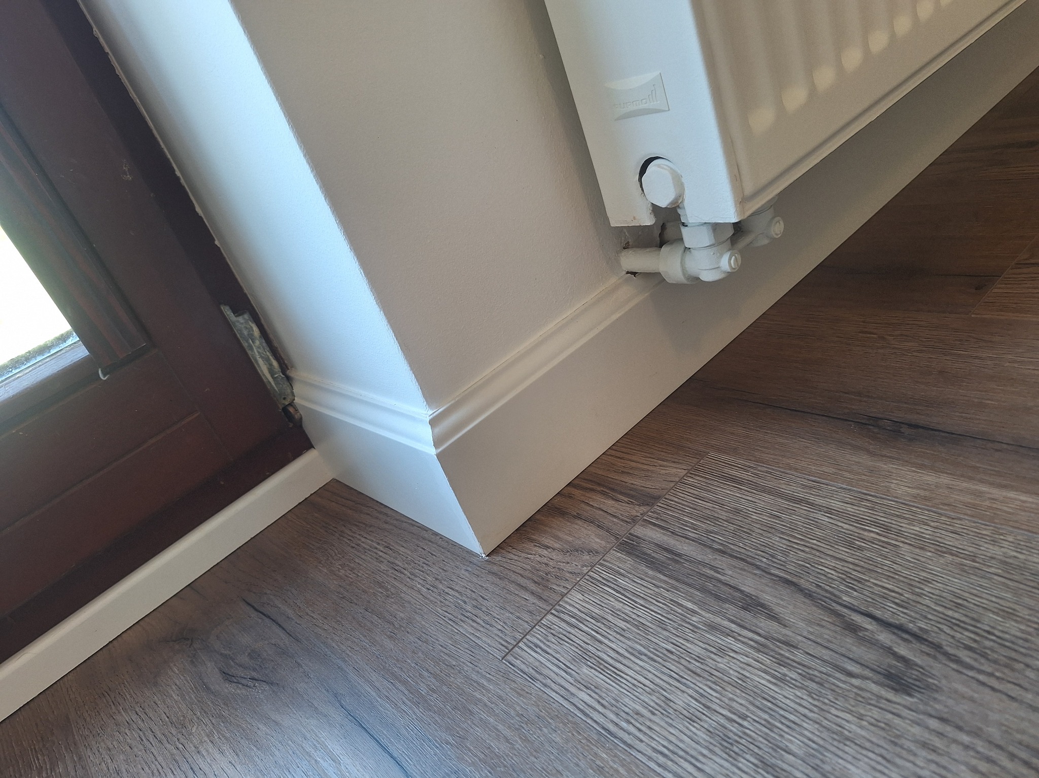 Biała listwa przypodłogowa z frezem, brązowe panele podłogowe i fragment grzejnika. Ujęcie z niskiego kąta, blisko okna.