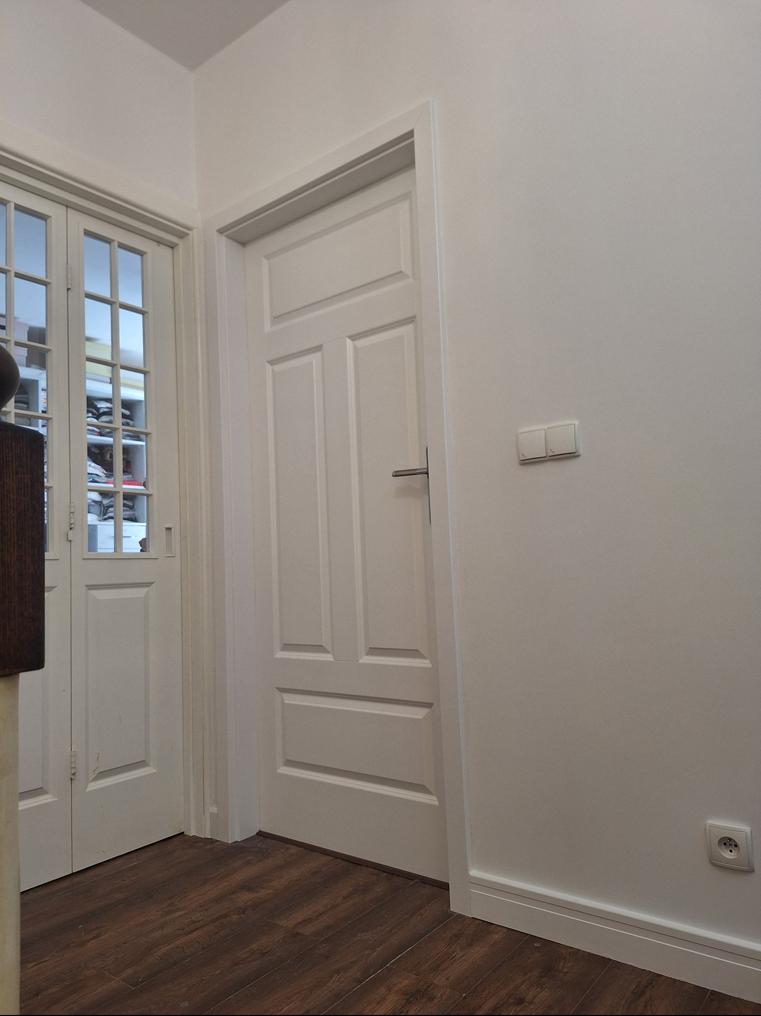 Jasne wnętrze z dwiema parami białych drzwi: pełne i przeszklone. Podłoga z paneli w odcieniach brązu. Minimalistyczny design, gładkie, białe ściany.