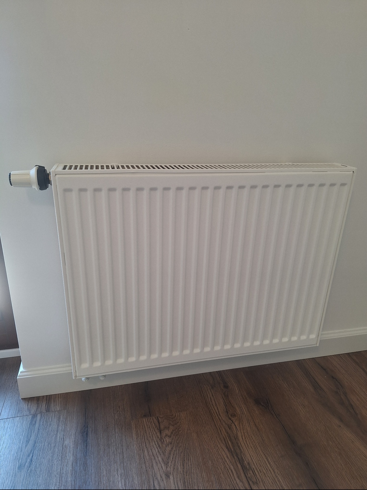 Biały, nowy grzejnik panelowy z termostatem zamontowany na ścianie, widoczny fragment drewnopodobnej podłogi i listwy przypodłogowej.