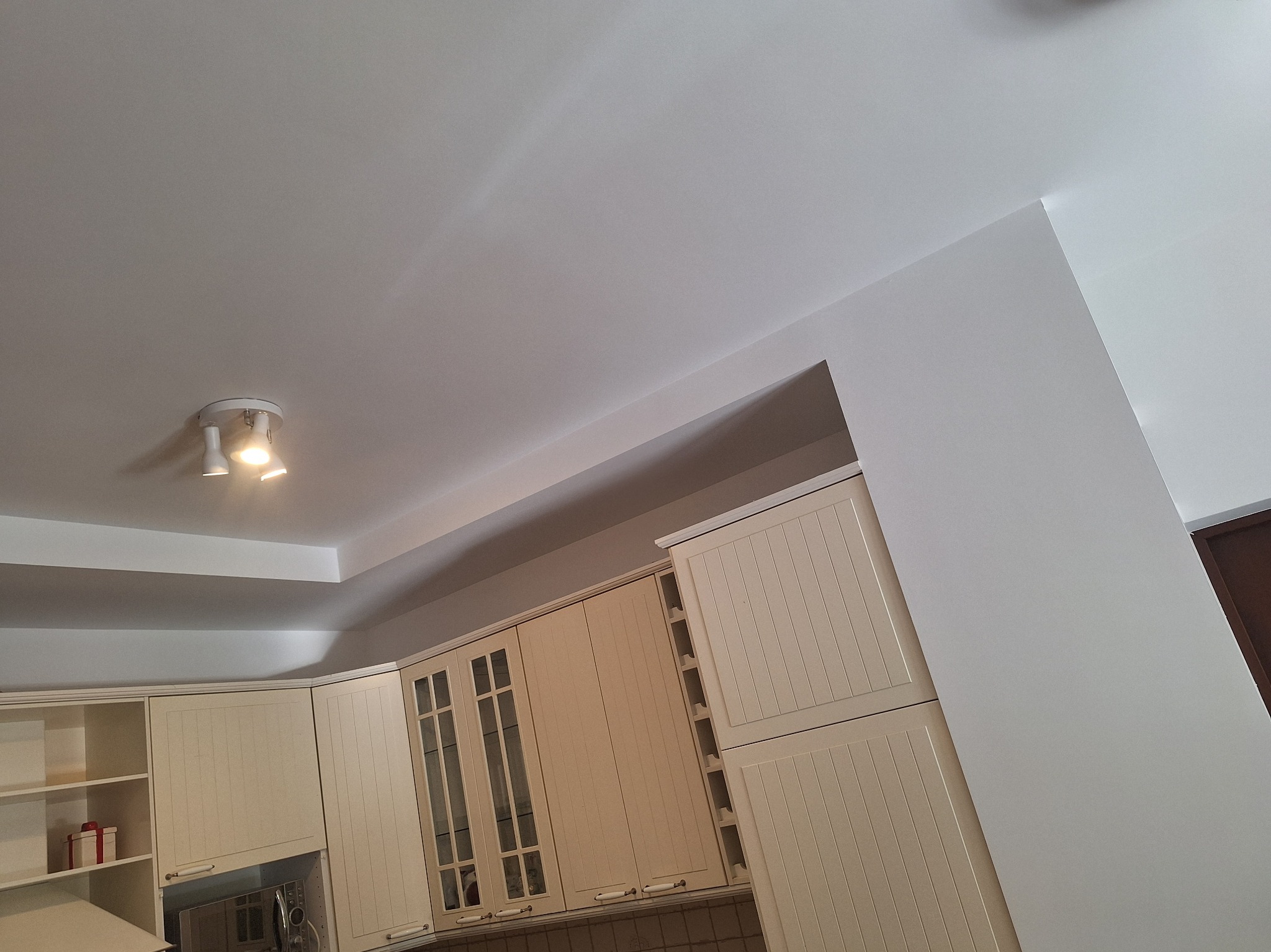 Jasna kuchnia z kremowymi szafkami pod sufitem i oświetleniem punktowym. Świeżo pomalowane ściany w odcieniach bieli. Widok na wnętrze.