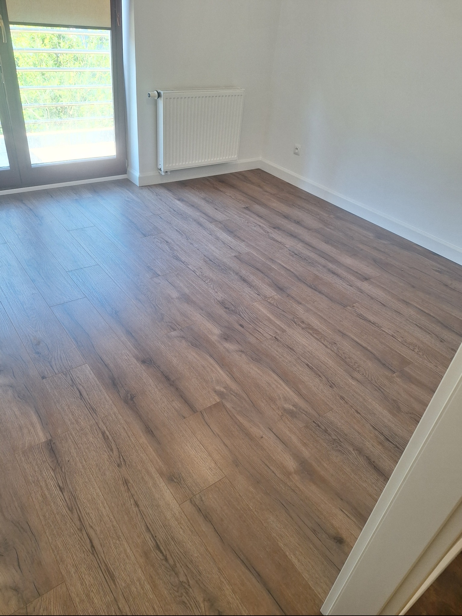 Jasne panele podłogowe z widoczną strukturą drewna w pustym pokoju z białymi ścianami i grzejnikiem. Okno balkonowe z widokiem na zieleń.