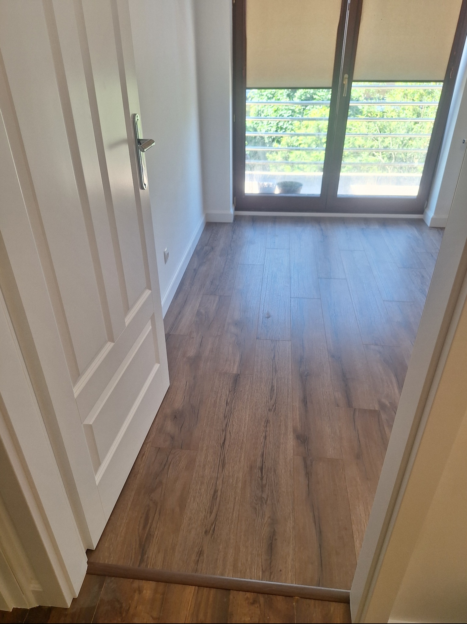 Wykończone wnętrze: brązowe panele podłogowe z widokiem na okno z roletami i częściowo otwarte białe drzwi z metalową klamką. Świeżo po remoncie.