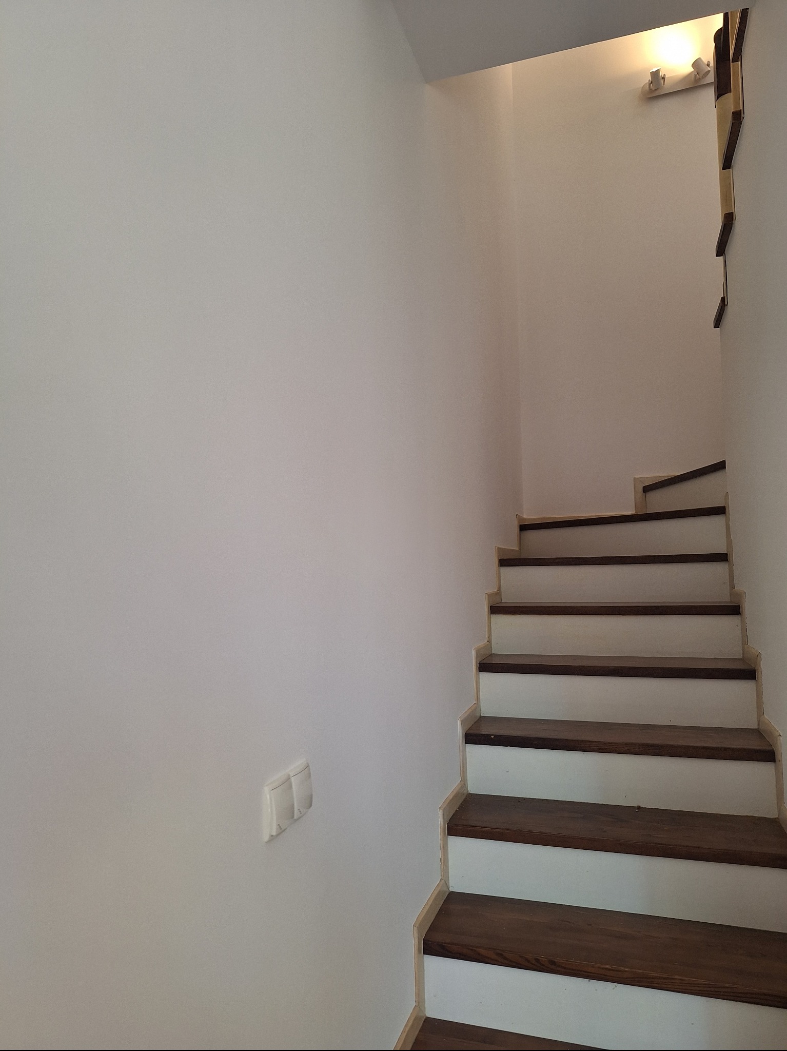 Białe schody z drewnianymi stopniami w jasnym wnętrzu. Ściany pomalowane na biało, widoczny włącznik światła i oświetlenie schodów.