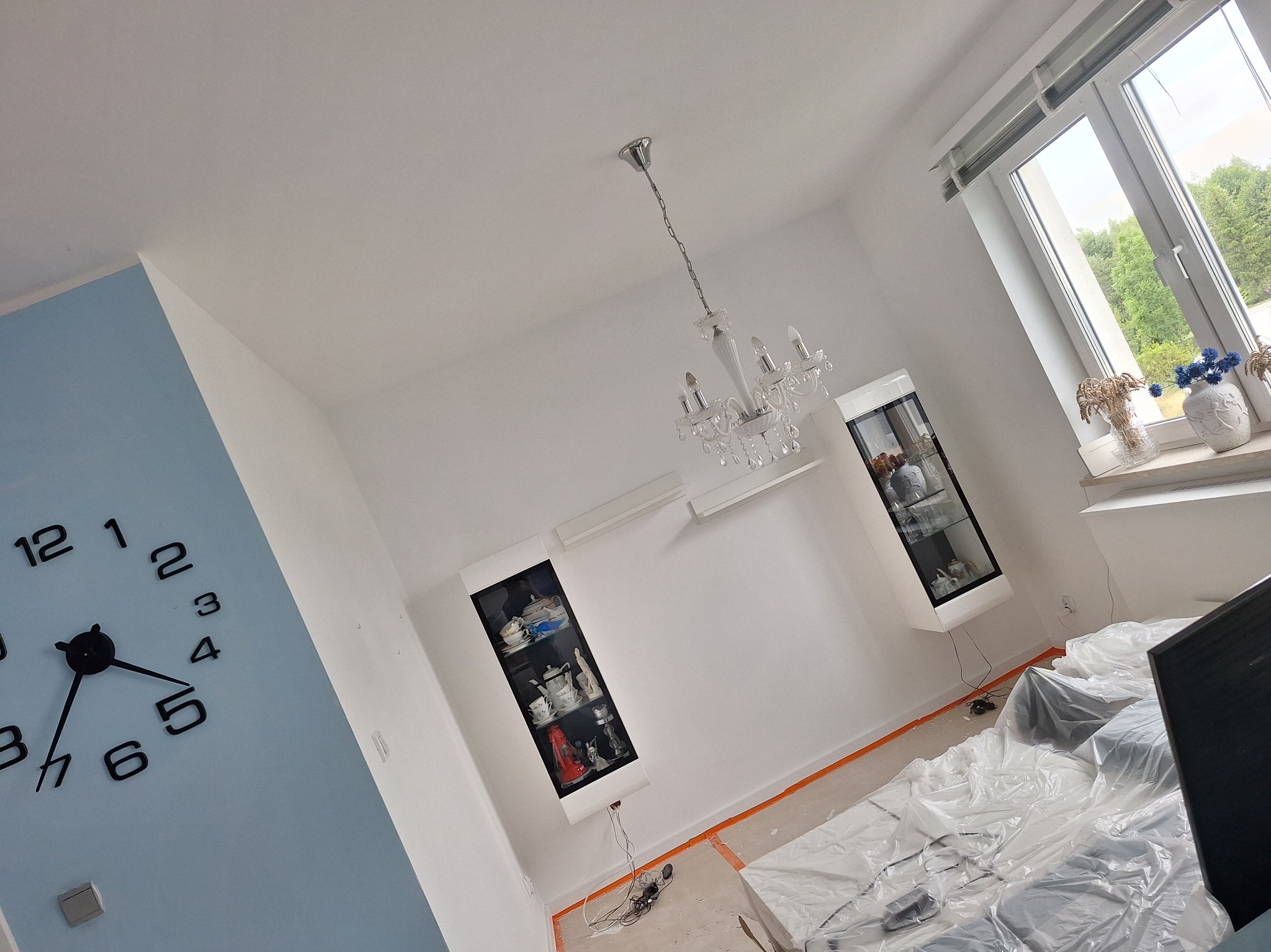 Jasne wnętrze w trakcie zabezpieczania folią malarską, z widocznymi detalami jak wiszący kryształowy żyrandol i nowoczesne szafki wiszące. Ściana w odcieniu błękitu z zegarem.