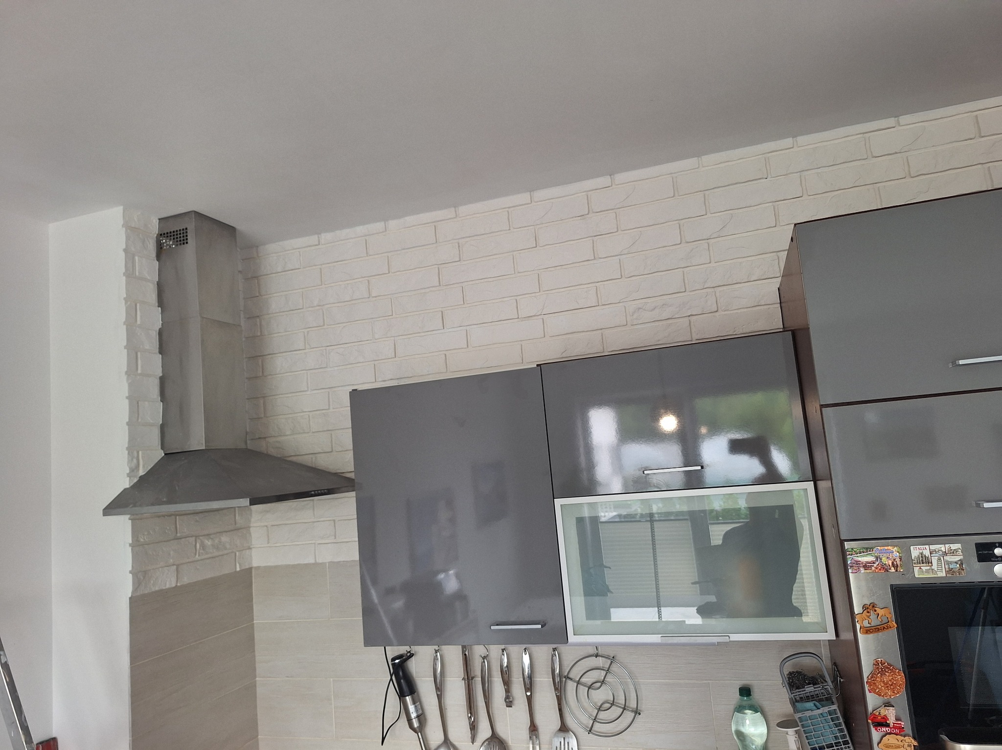 Wykończona kuchnia z szarymi szafkami, białymi cegiełkami na ścianie i srebrnym okapem. Widoczne akcesoria kuchenne i odbicie w szafkach.