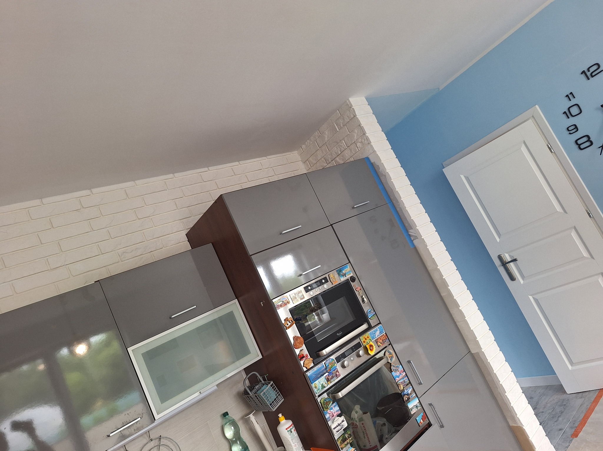 Nowoczesna kuchnia z szarymi szafkami i białymi cegiełkami na ścianie, widok z góry, po remoncie. Na ścianie niebieska farba i białe drzwi z zegarem.