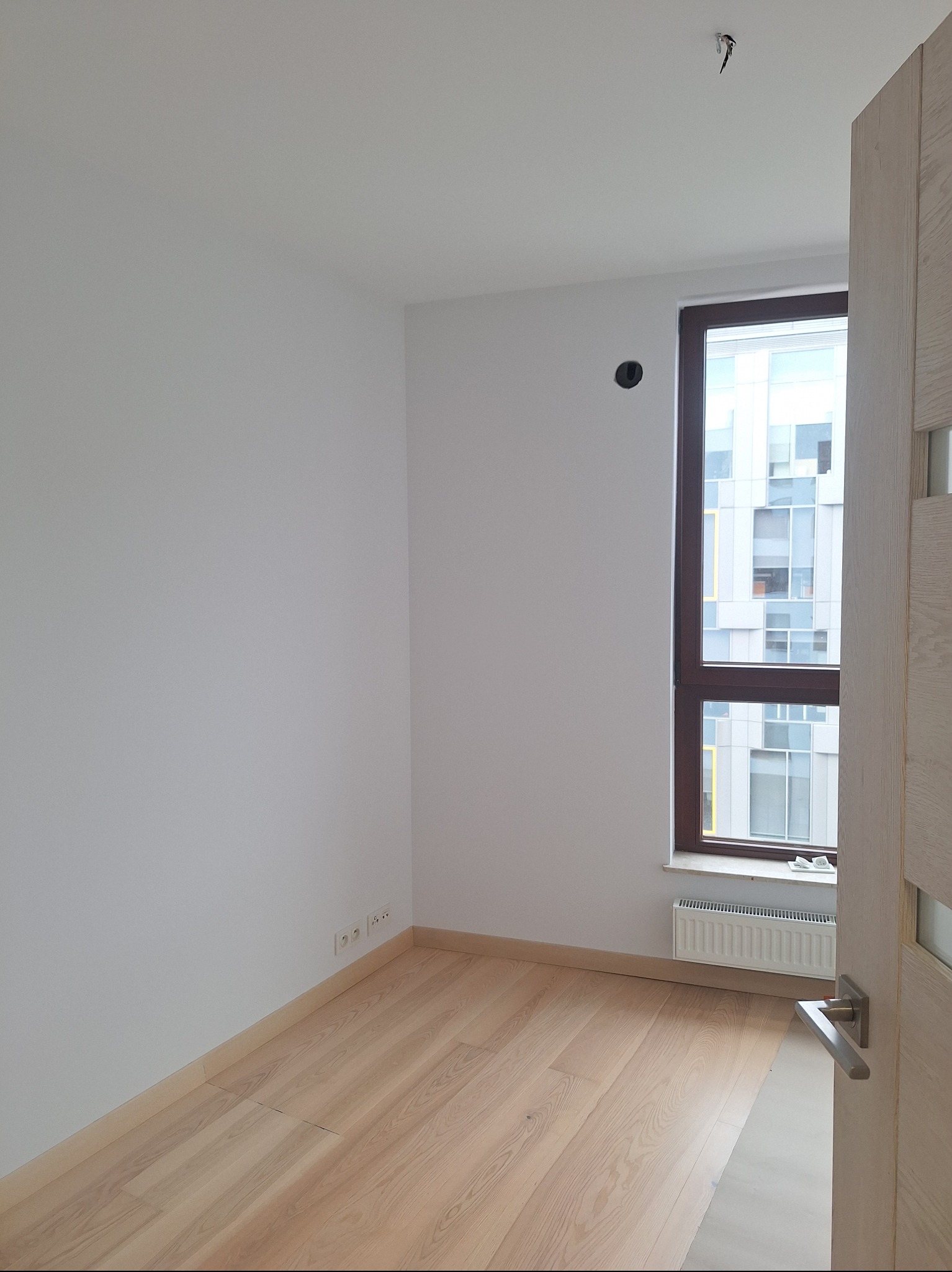 Jasne, puste wnętrze z panelami podłogowymi i białymi ścianami. Widoczne gniazdka elektryczne przy podłodze, okno z grzejnikiem i fragment otwartych drzwi.