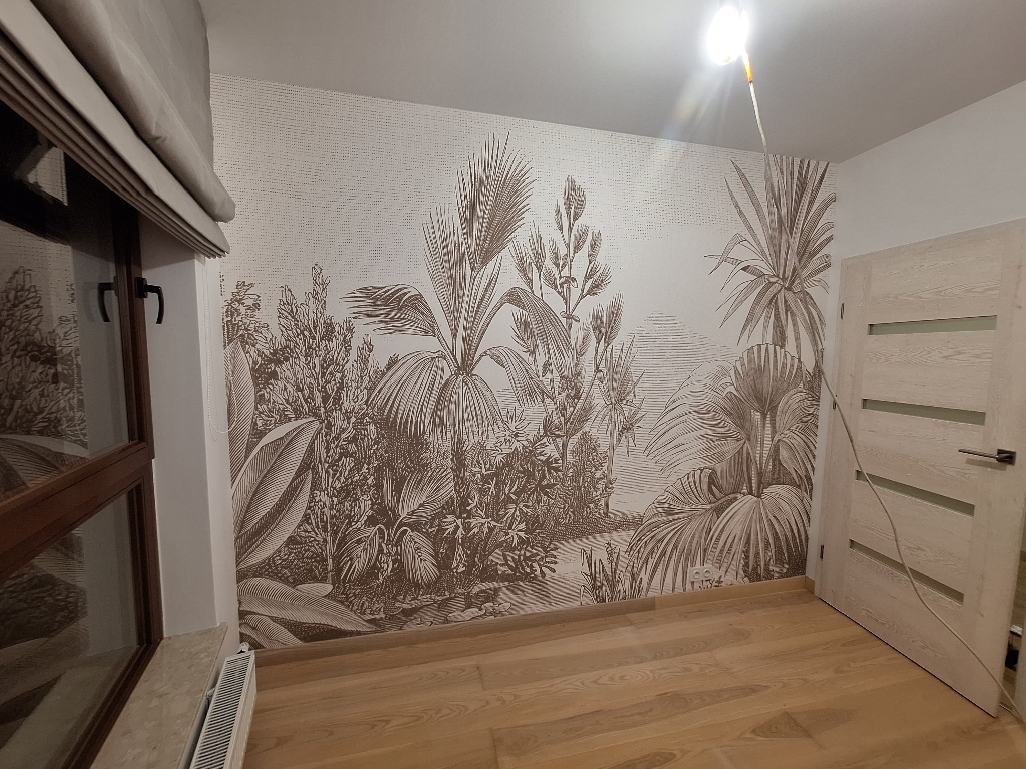 Pokój po tapetowaniu z motywem roślinnym w stylu vintage. Jasne panele podłogowe, okno z roletą rzymską i białe drzwi. Widoczny fragment grzejnika.