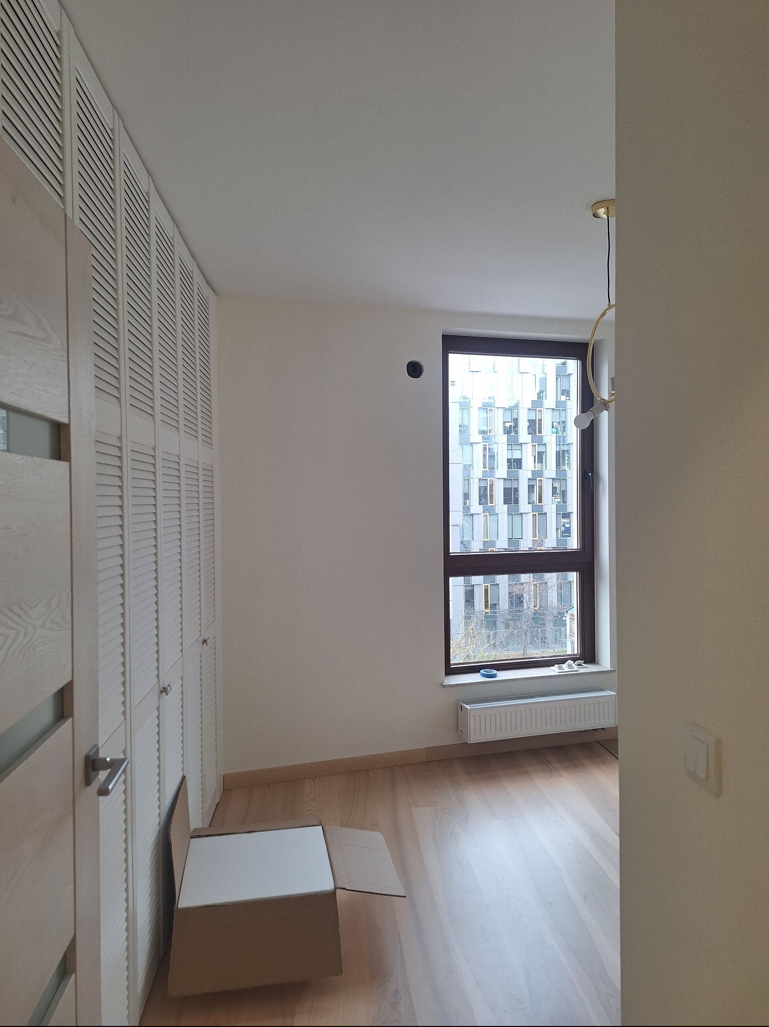 Jasne wnętrze pokoju z drewnianą podłogą i oknem z widokiem na budynek. Przy ścianie szafa z lamelowymi drzwiami. Na podłodze kartonowe pudło.