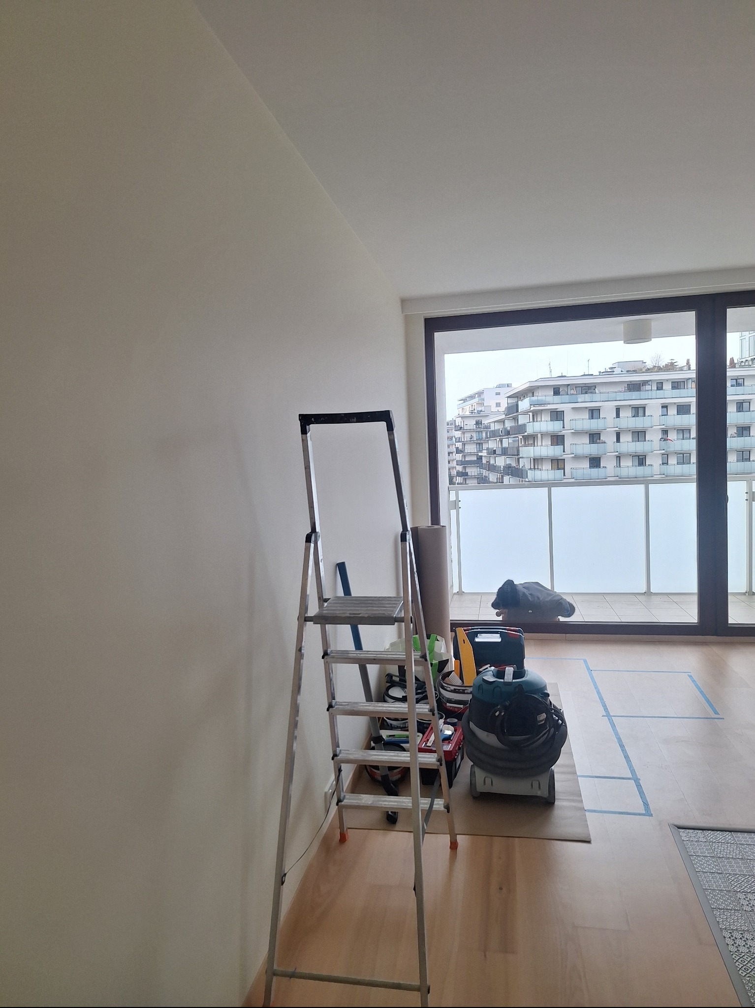 Przygotowania do malowania: drabina, narzędzia i odkurzacz na podłodze zabezpieczonej taśmą malarską, ściana gotowa do malowania, widok na balkon i budynki.