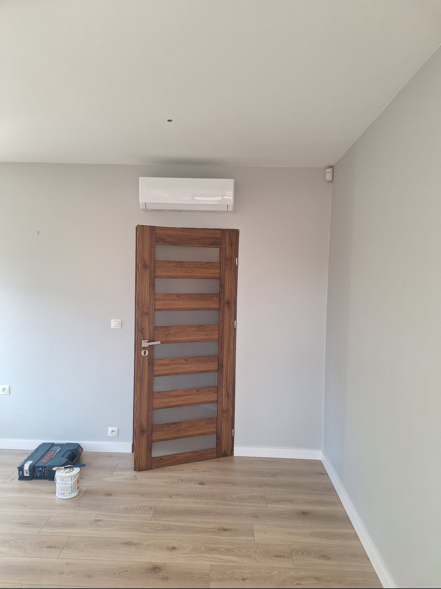 Wnętrze po remoncie w Piasecznie: drewniane drzwi z przeszkleniami, świeżo pomalowane szare ściany, jasna podłoga. Widoczny klimatyzator i akcesoria malarskie.