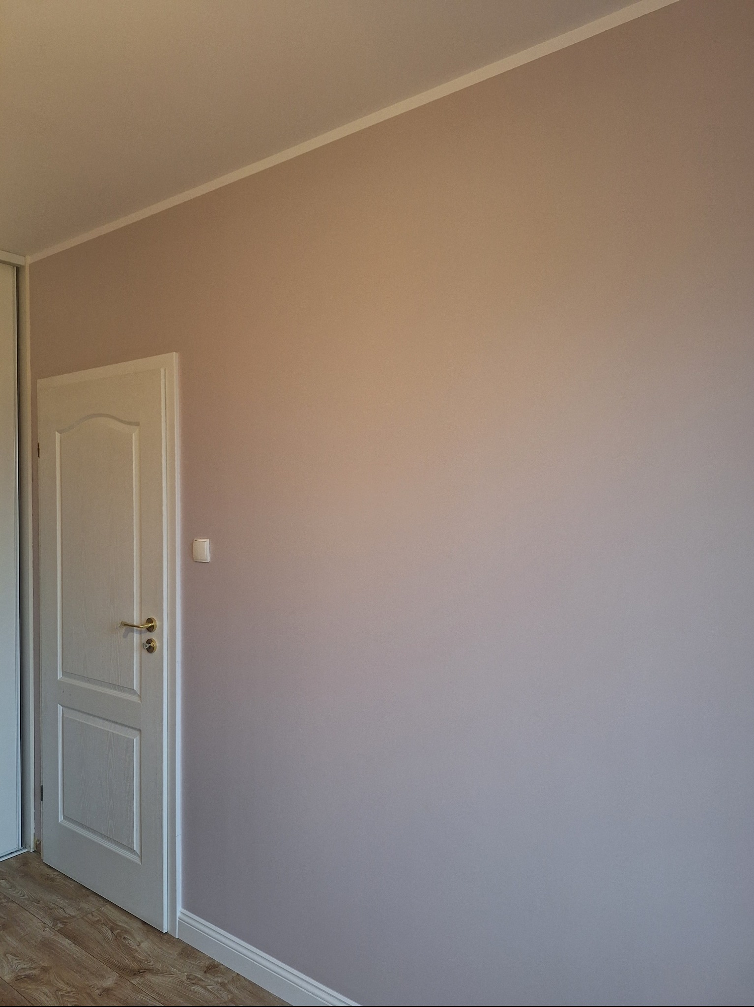 Ściana w odcieniach beżu i fioletu po malowaniu, widoczna biała listwa przypodłogowa i drzwi z ozdobnymi frezami. Delikatne światło podkreśla gładką powierzchnię.