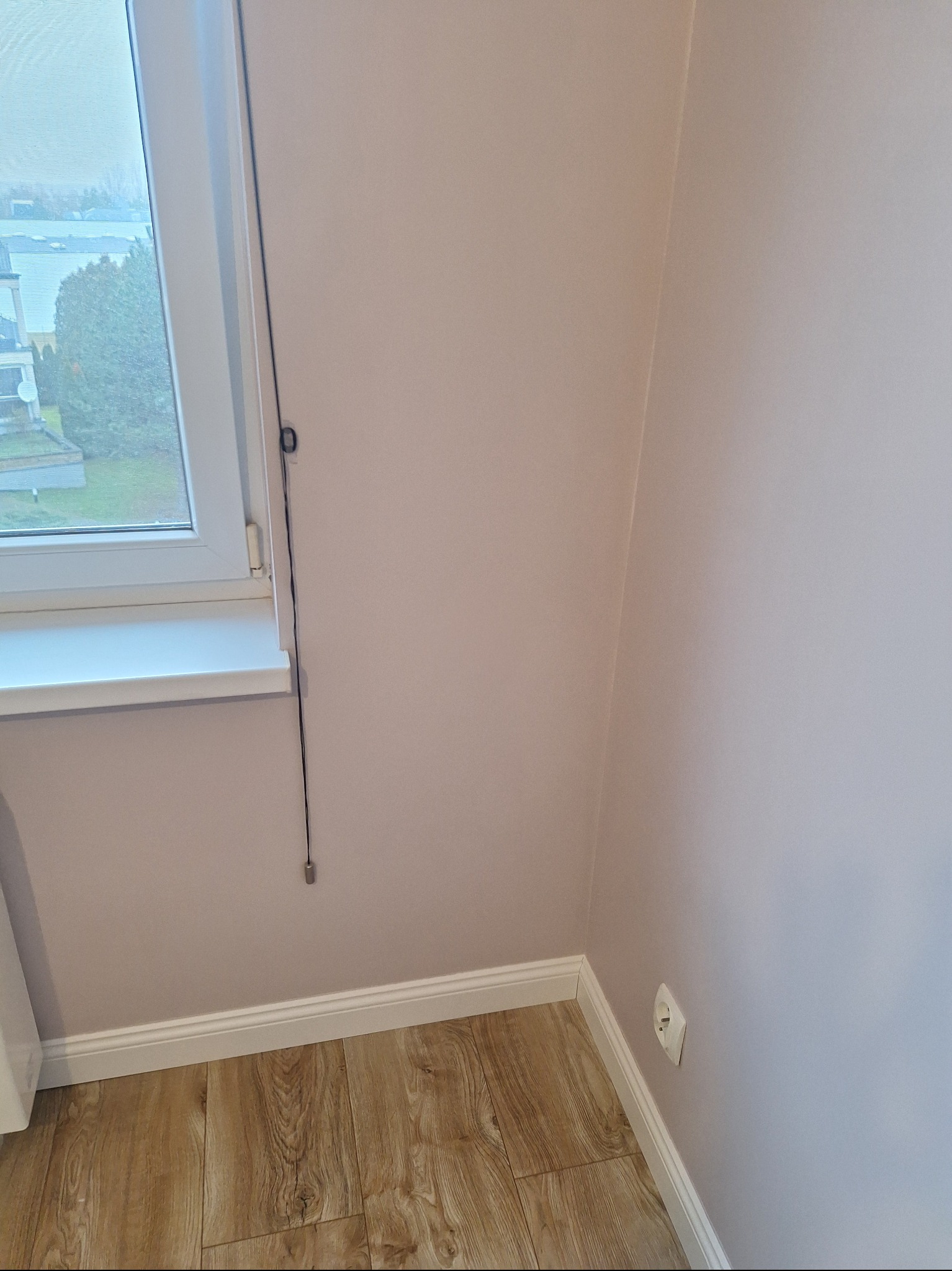 Jasny pokój po remoncie: panele podłogowe, pomalowane ściany w odcieniach beżu, biała listwa przypodłogowa i okno z widokiem na zieleń. Gniazdko elektryczne na ścianie.