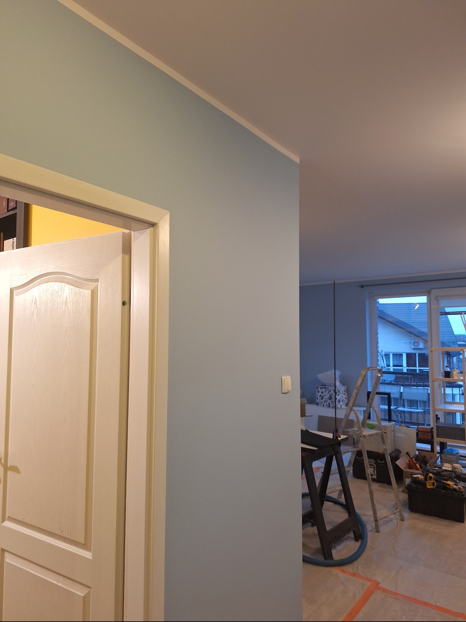 Świeżo pomalowana na błękitno ściana z białą listwą przypodłogową, widoczny fragment białych drzwi i drabina w tle. Wnętrze w trakcie prac wykończeniowych.