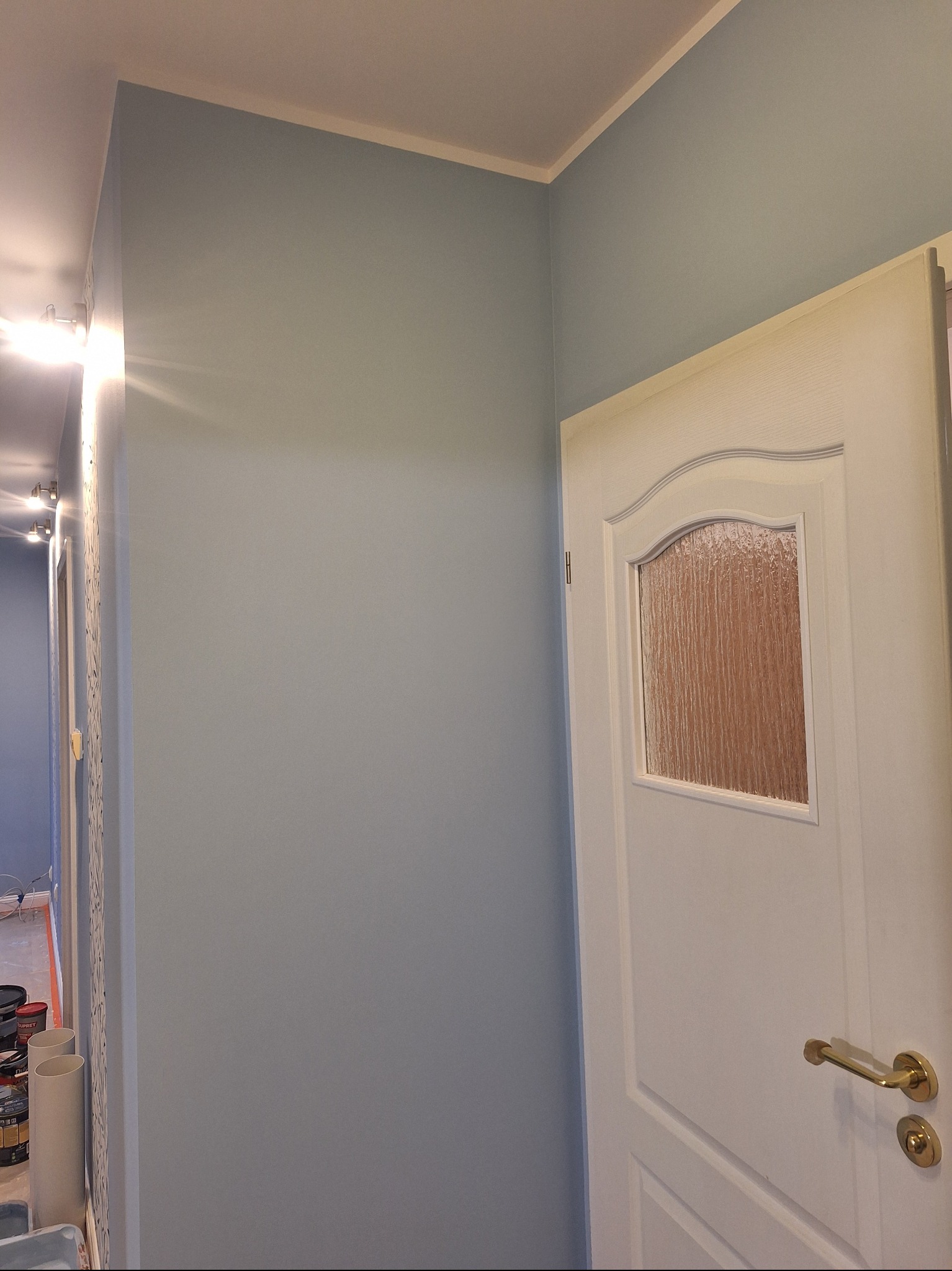 Ściana pomalowana na błękitno z białą, częściowo otwartą, zdobioną  drzwią z szybą. Widoczne puszki z farbą w tle sugerują świeżo zakończone prace.