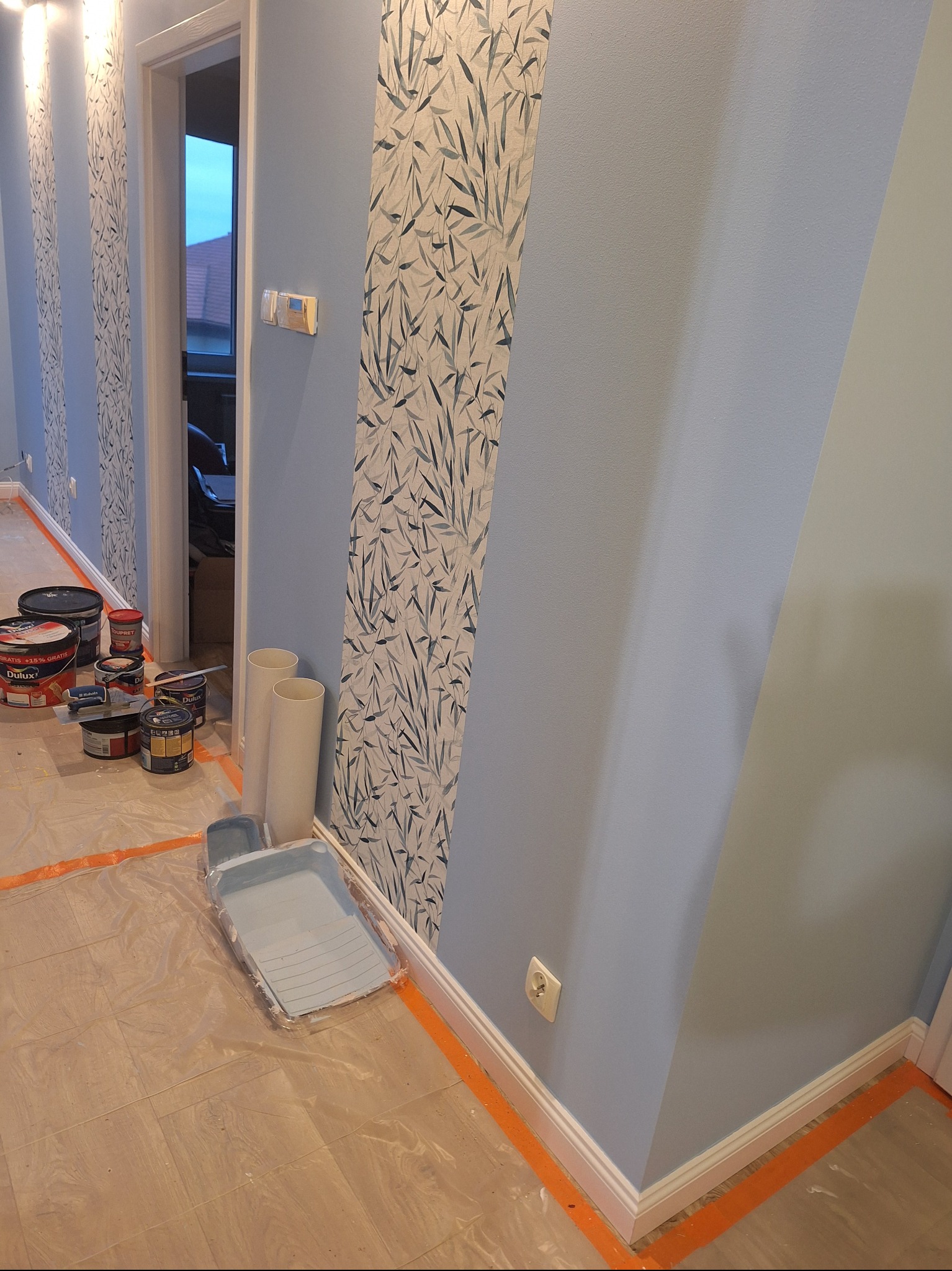 Pomalowane na niebiesko ściany z dekoracyjnym pasem z motywem liści. Widoczne puszki z farbą, wałki i kuweta malarska. Podłoga zabezpieczona folią malarską.