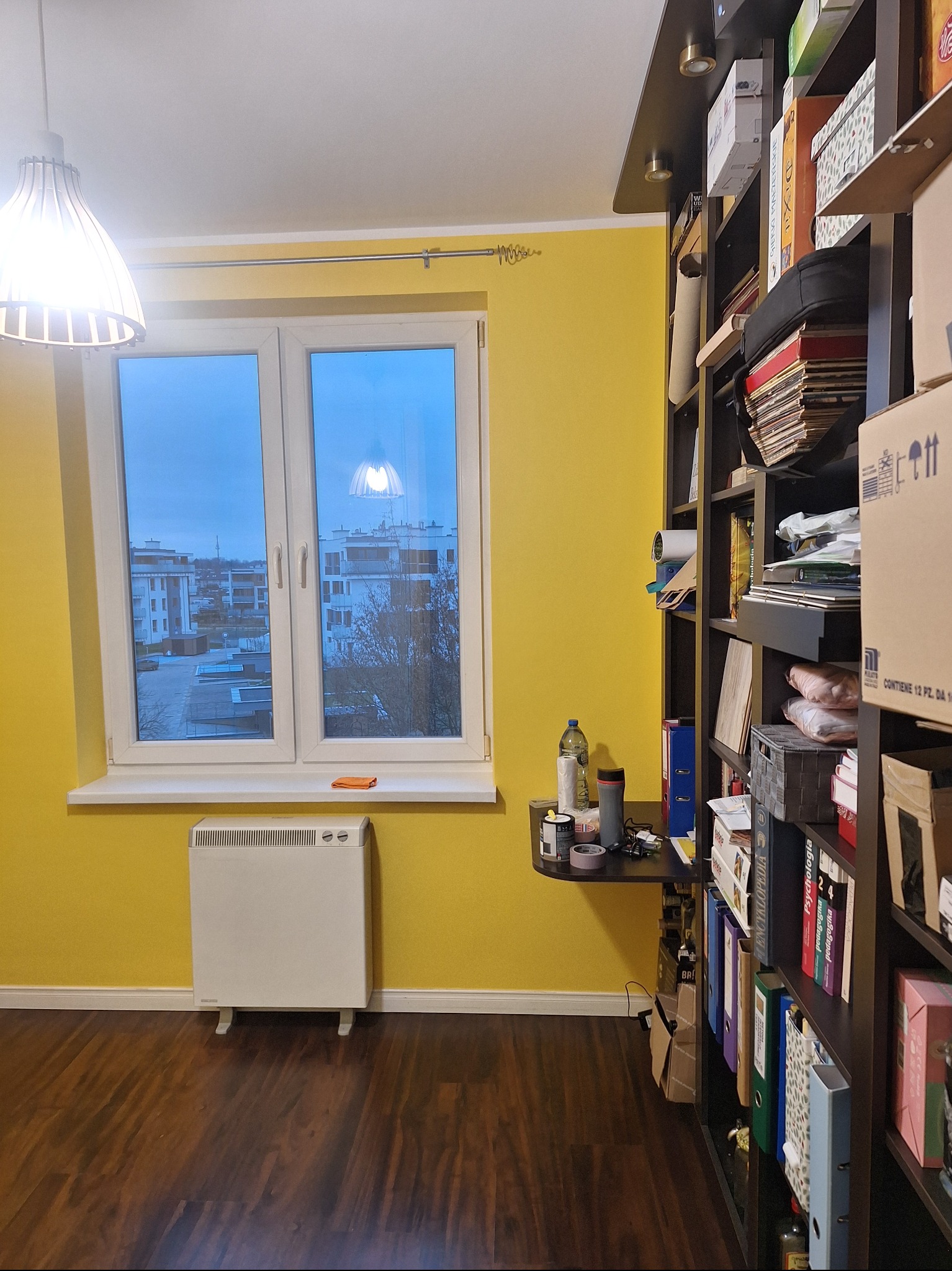 Żółty pokój z drewnianą podłogą, białym grzejnikiem i regałem pełnym książek. Widok z okna na budynki. Lampa sufitowa w stylu skandynawskim.