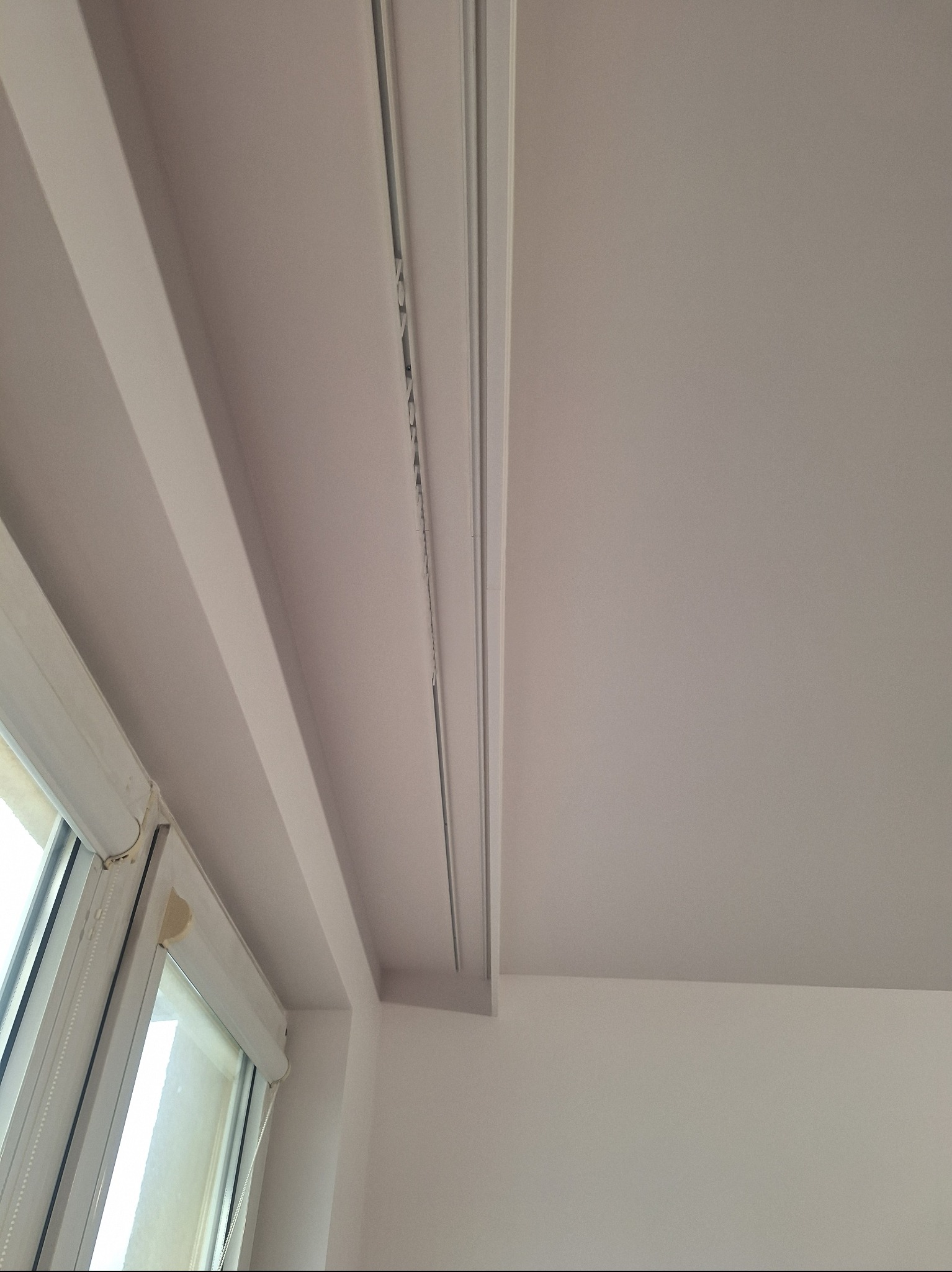 Białe, gładkie wykończenie sufitu z zamontowaną szyną karniszową, widoczne okno z roletą. Minimalistyczny design wnętrza, neutralna kolorystyka.