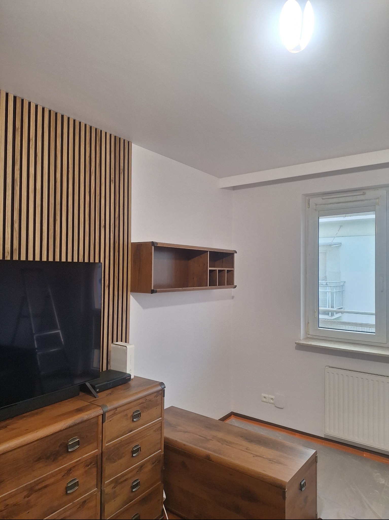 Jasne wnętrze pokoju po remoncie. Na ścianie drewniane lamele, podwieszana półka i okno. Na podłodze zabezpieczona folią podłoga. Widoczny drewniany komoda i skrzynia.