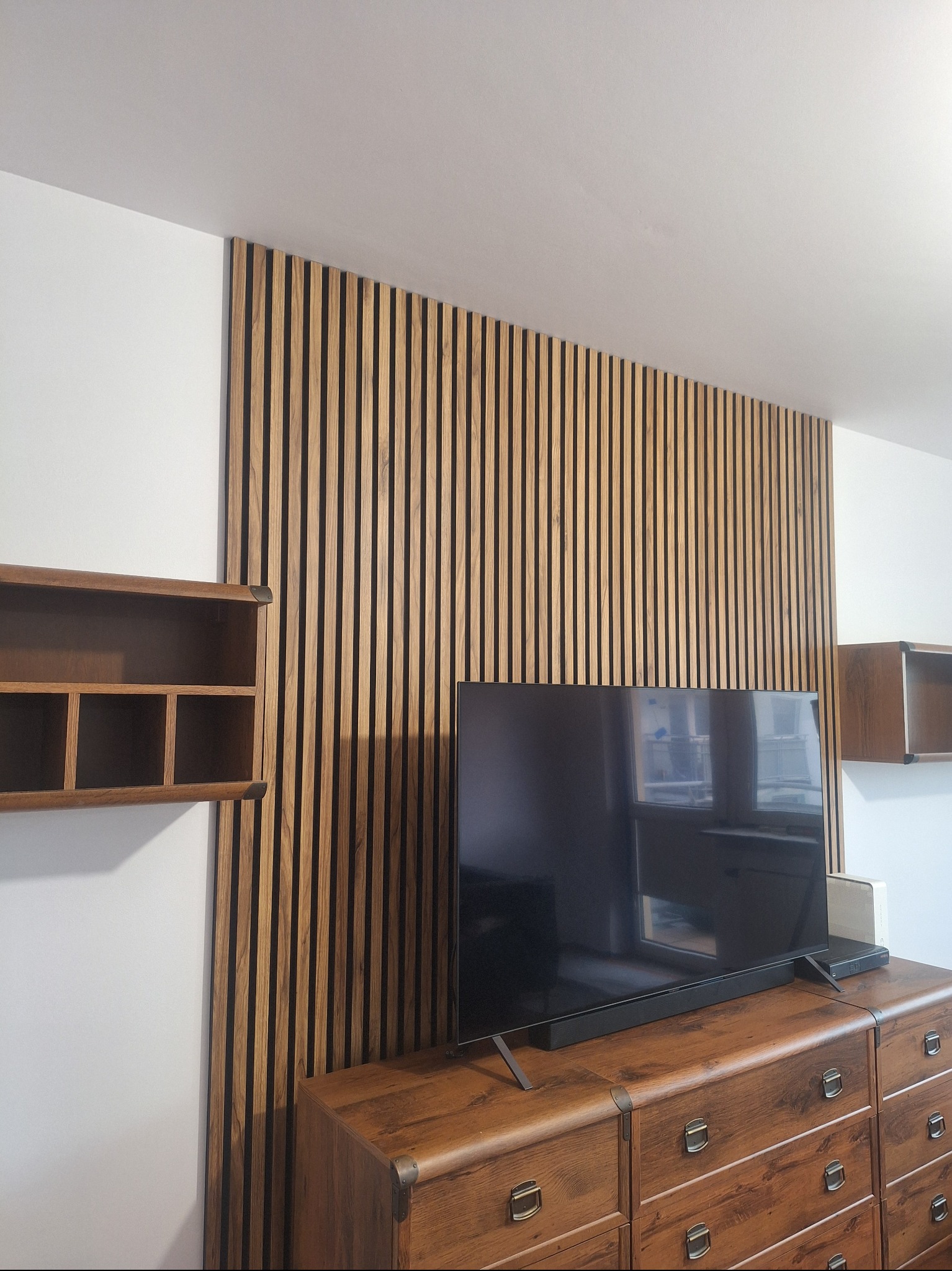 Drewniana ściana dekoracyjna z lameli w salonie w Piasecznie. Telewizor na komodzie, półki wiszące. Nowoczesny design wnętrza z elementami drewna.