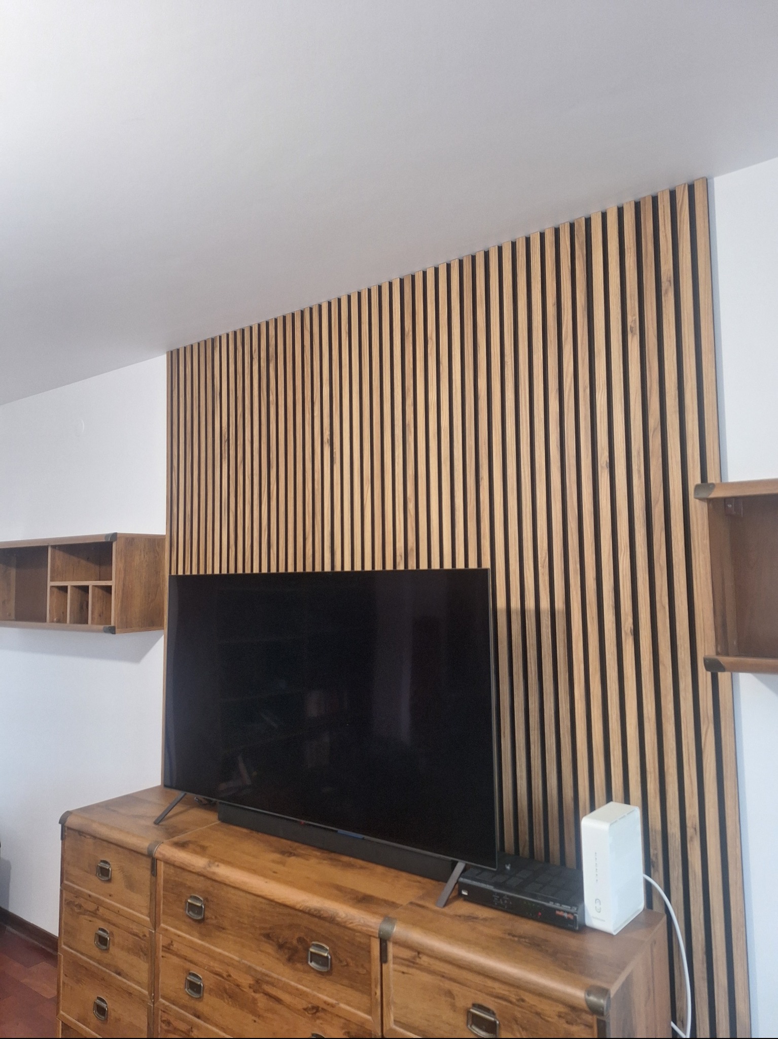 Drewniane lamele na ścianie za telewizorem, komoda z szufladami w stylu vintage, półka wisząca. Aranżacja wnętrza z elementami drewna.