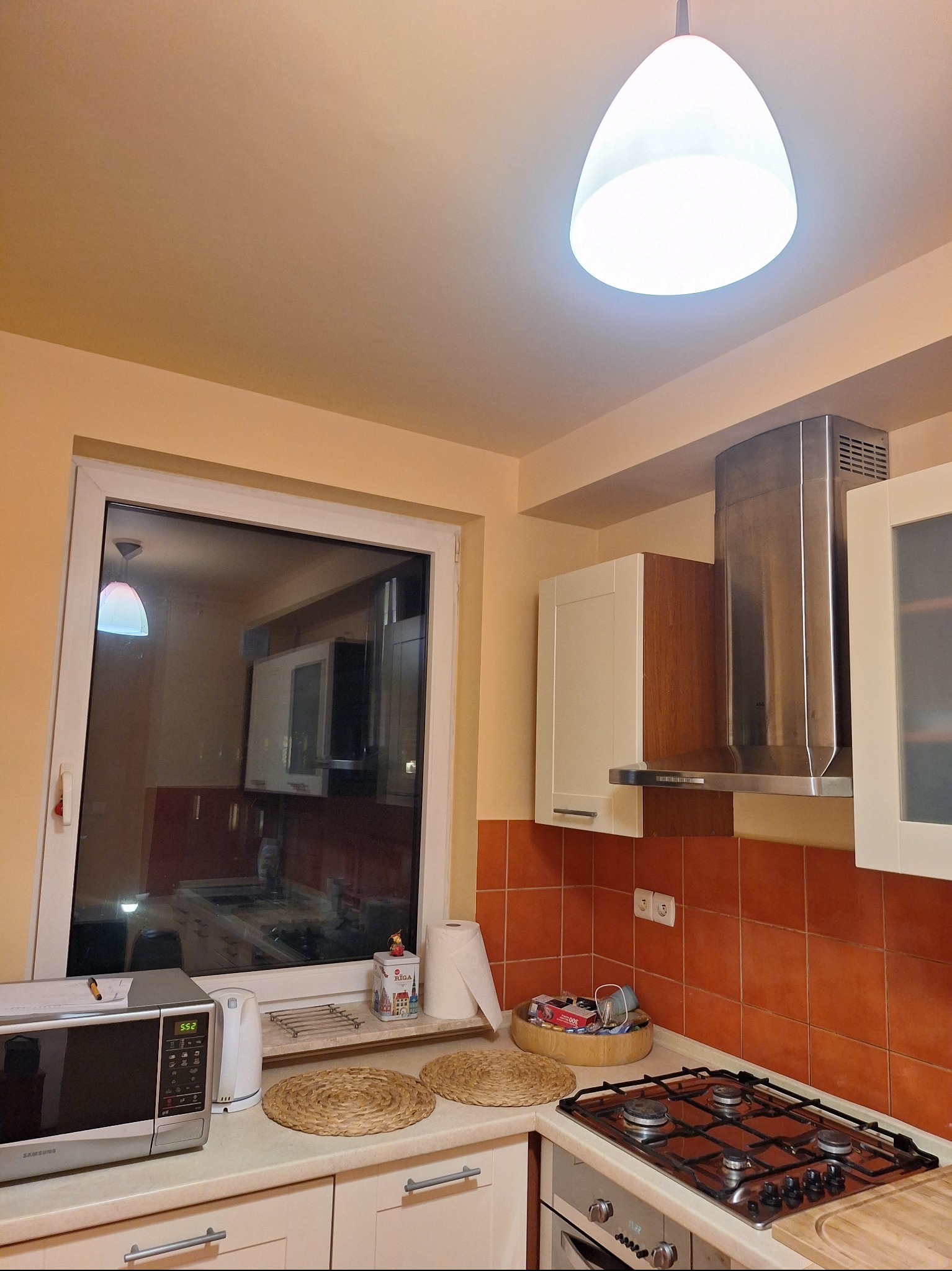 Jasna kuchnia z pomarańczowymi płytkami, widok na okno i sprzęty AGD. Ujęcie z perspektywy sufitu, z lampą w centrum kadru. Widoczne detale wyposażenia.
