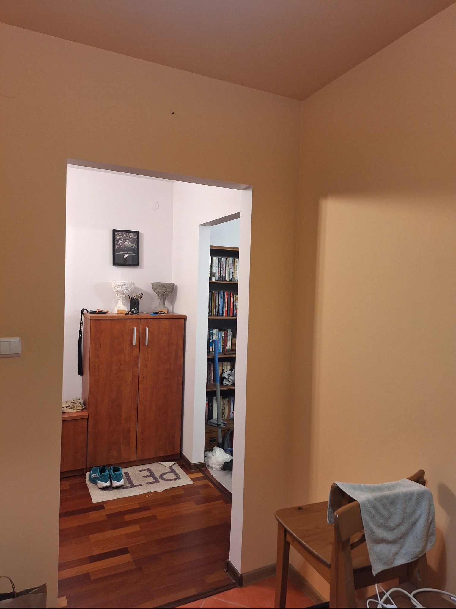 Korytarz po odświeżeniu: ściany w odcieniach beżu, drewniana podłoga, widok na szafkę z dekoracjami i regał z książkami. Krzesło z szarą szmatką w prawym dolnym rogu.