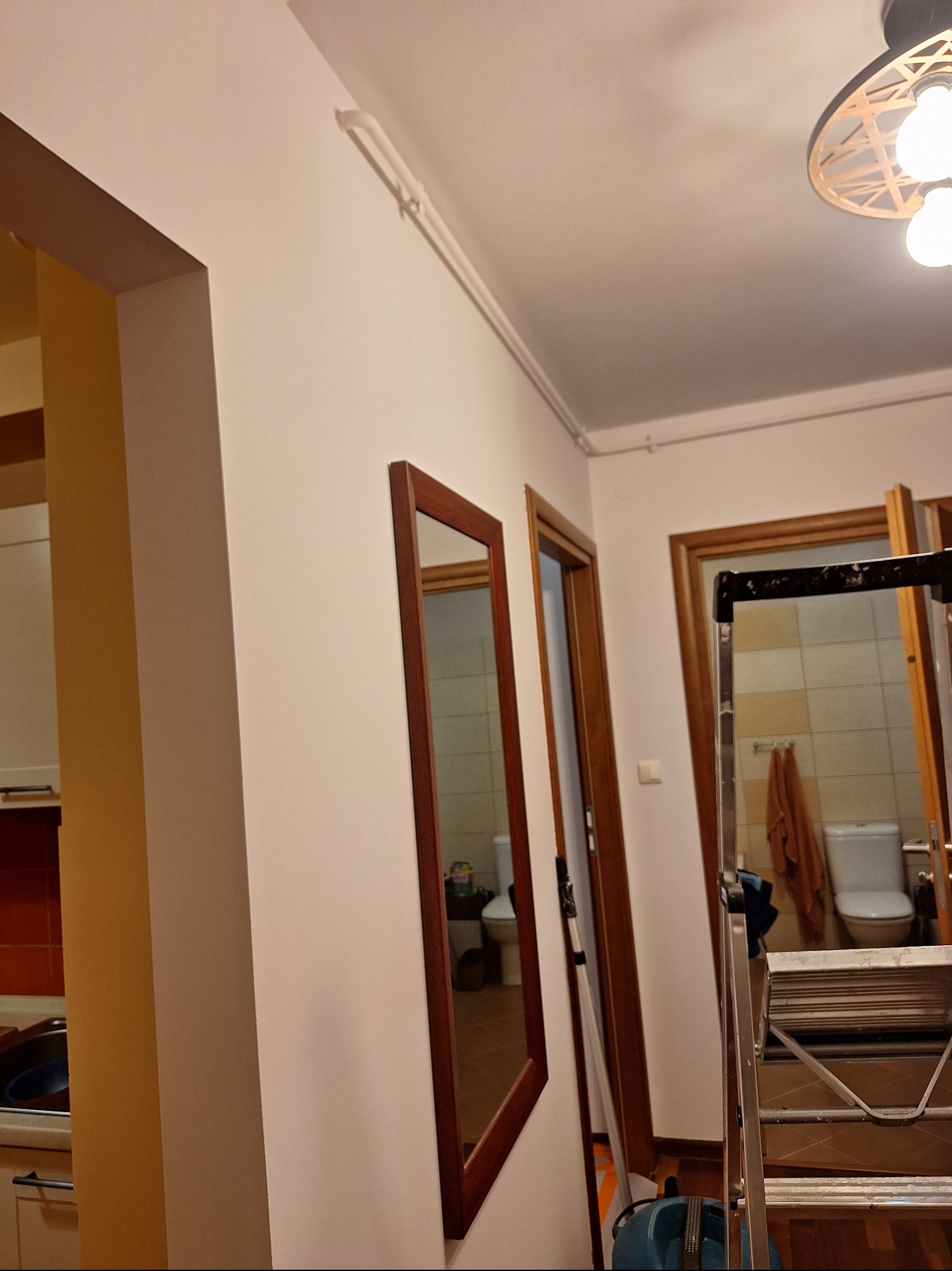 Biała ściana z lustrem w drewnianej ramie, widoczne rury pod sufitem i drabina. W tle widoczne drzwi do łazienki i kuchni.