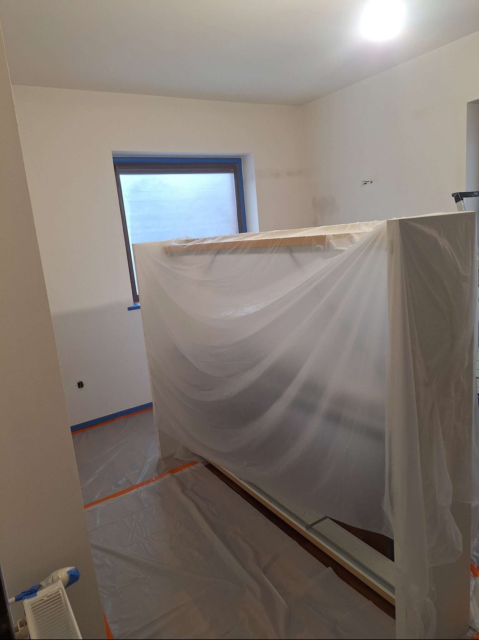 Pokój przygotowany do malowania: meble zabezpieczone folią, podłoga zakryta, okno z taśmą malarską. Widoczne świeżo pomalowane ściany w jasnym odcieniu.